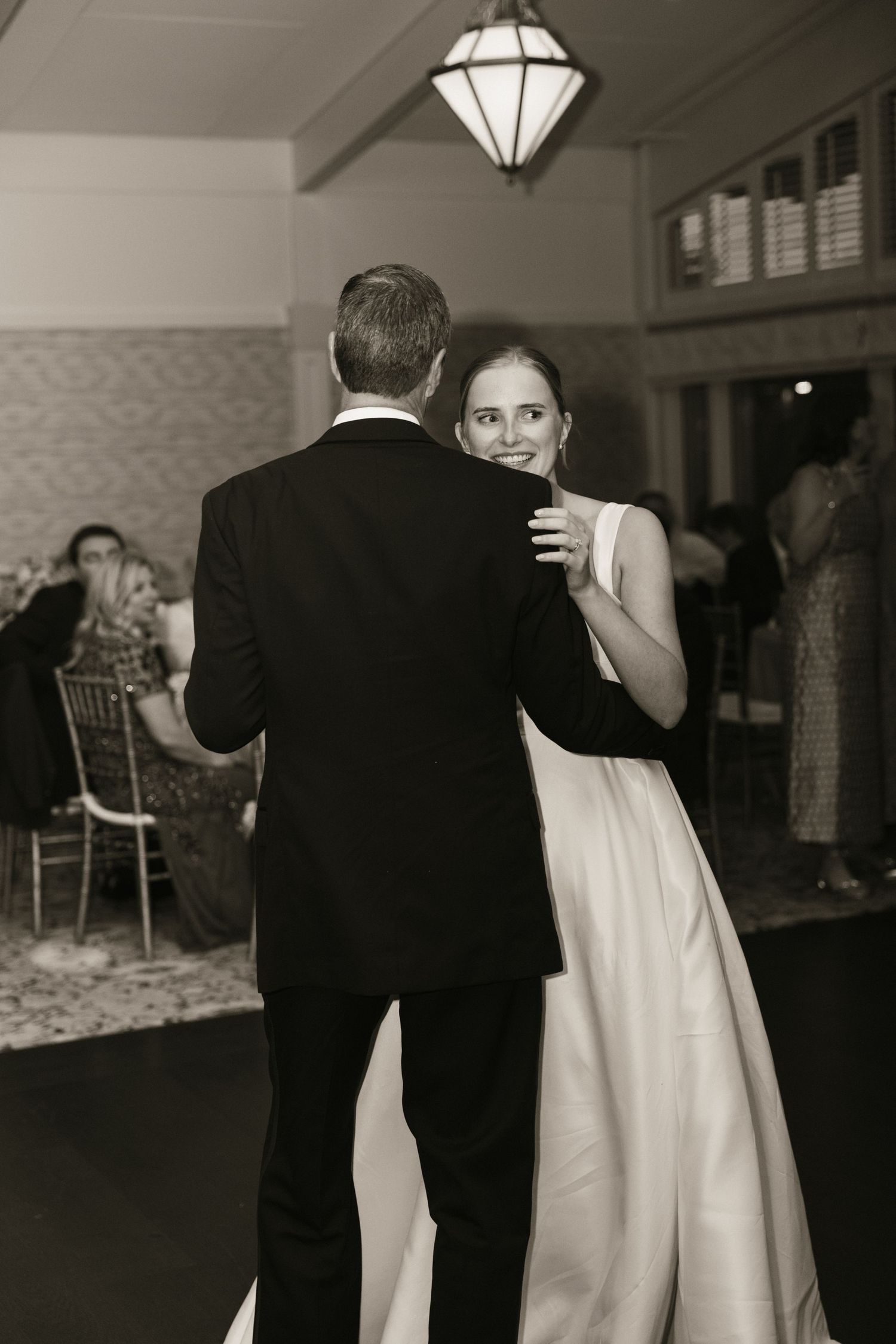 Black and white photo of bride smiling over grooms shoulder during romantic first dance at reception.