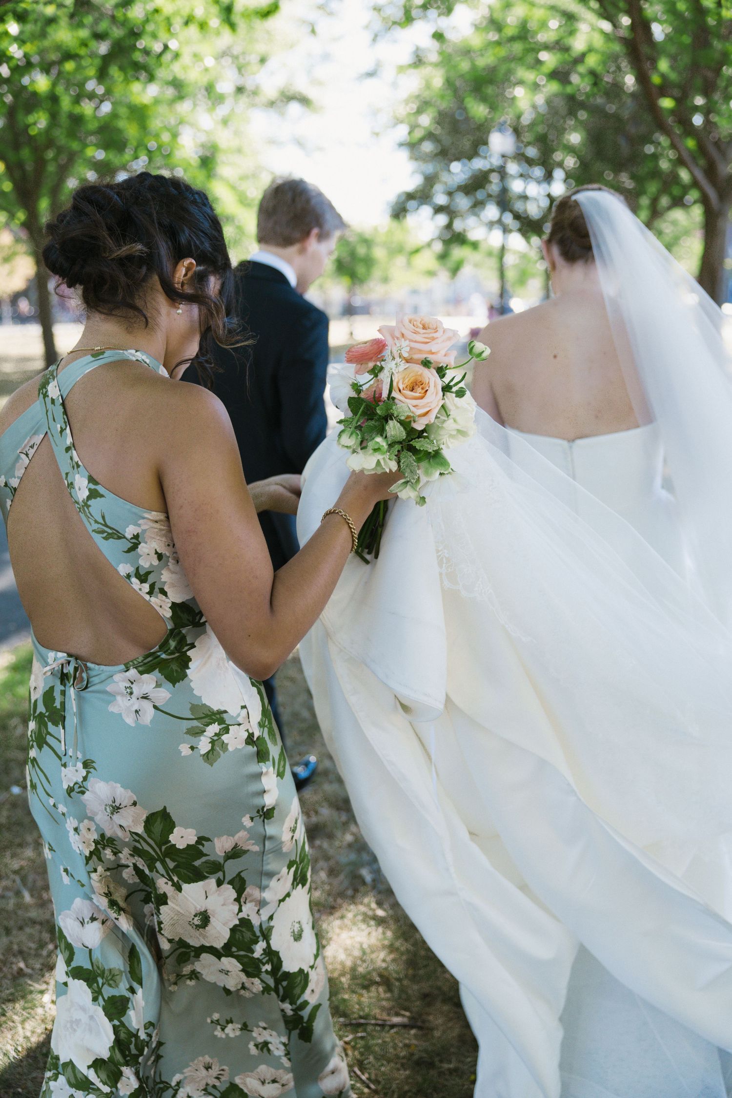Bridesmaid in floral dress helps bride with bouquet and veil outdoors before wedding ceremony.