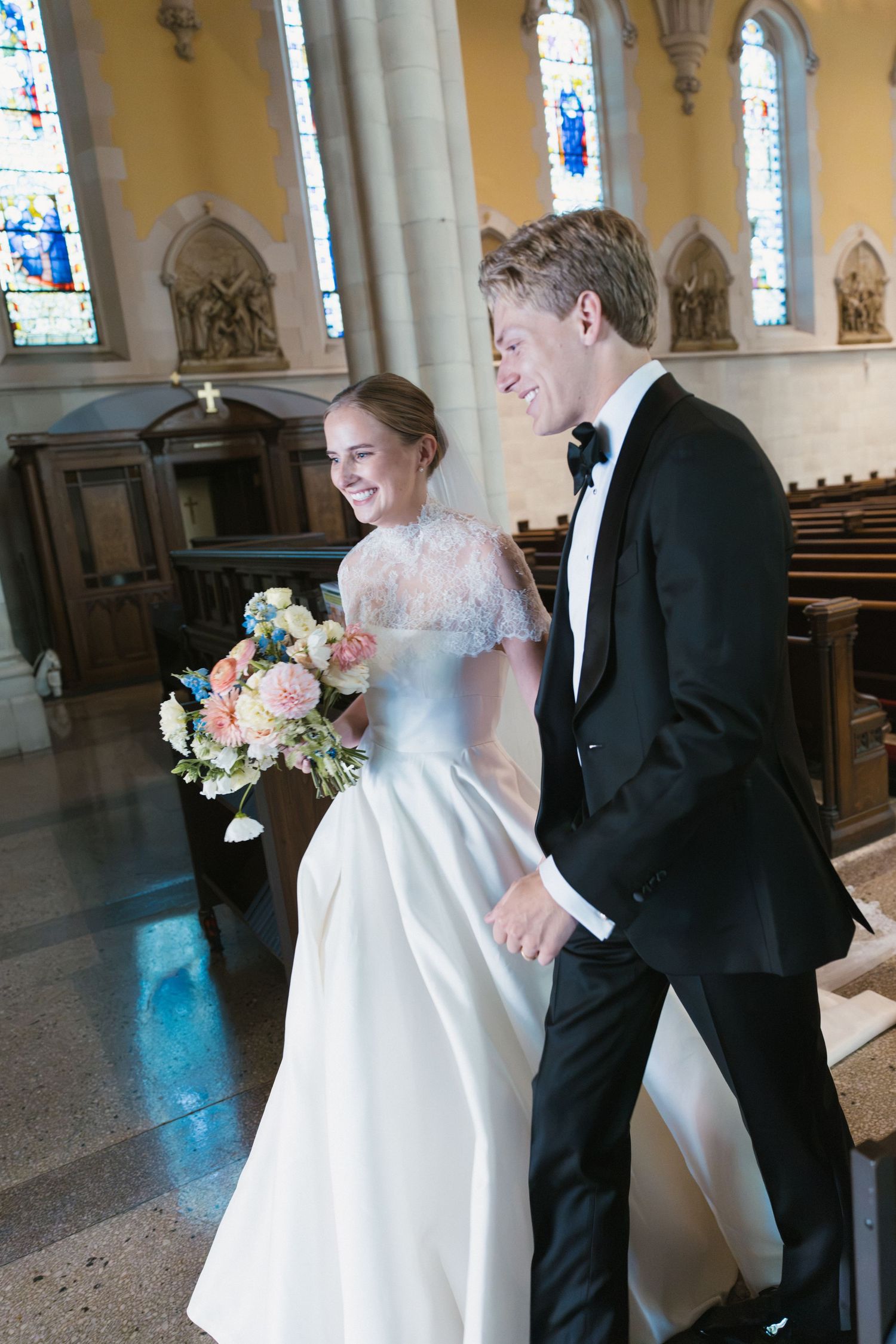 Smiling bride and groom walking down church aisle, bride holding colorful bouquet, groom in black tuxedo, stained glass windows.