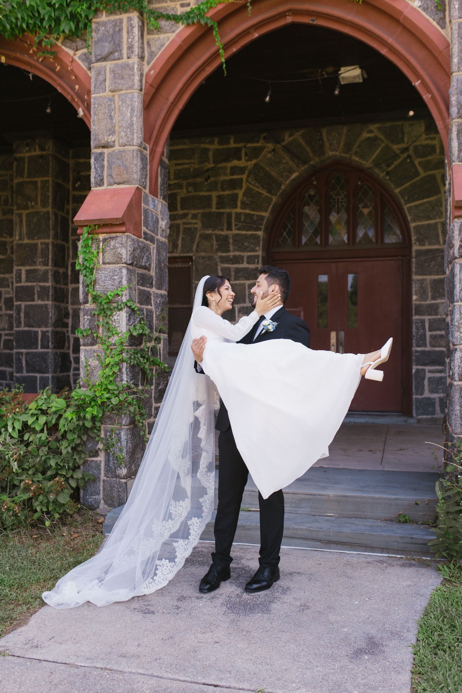 Groom carries bride in white lace veil outside stone church with arched wooden doors.