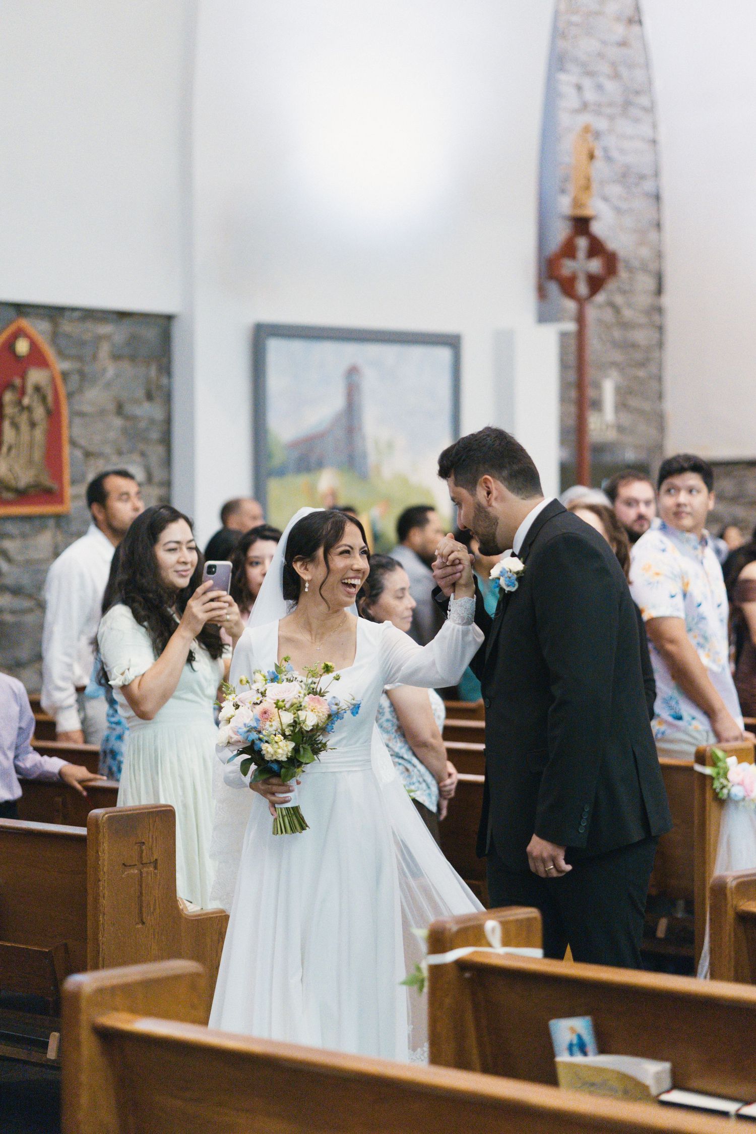 Bride and groom walk down church aisle laughing, as bridesmaid captures moment on phone during wedding ceremony.