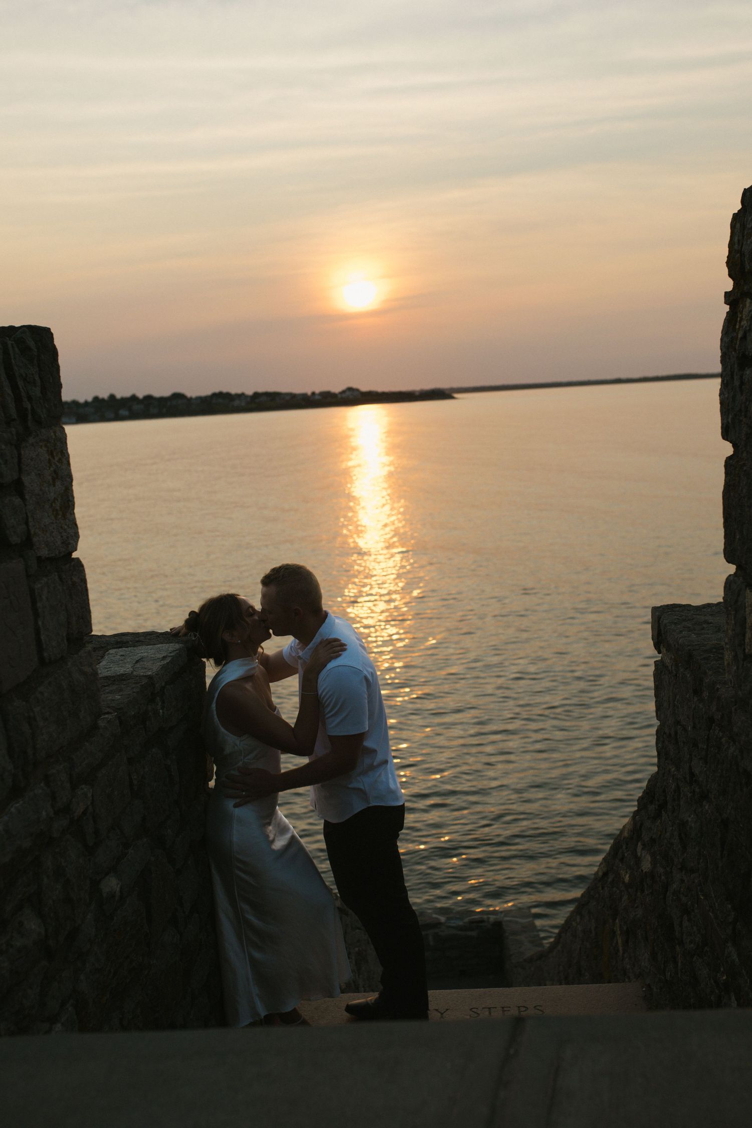 Couple sharing a romantic kiss silhouetted against a golden sunset over the water.