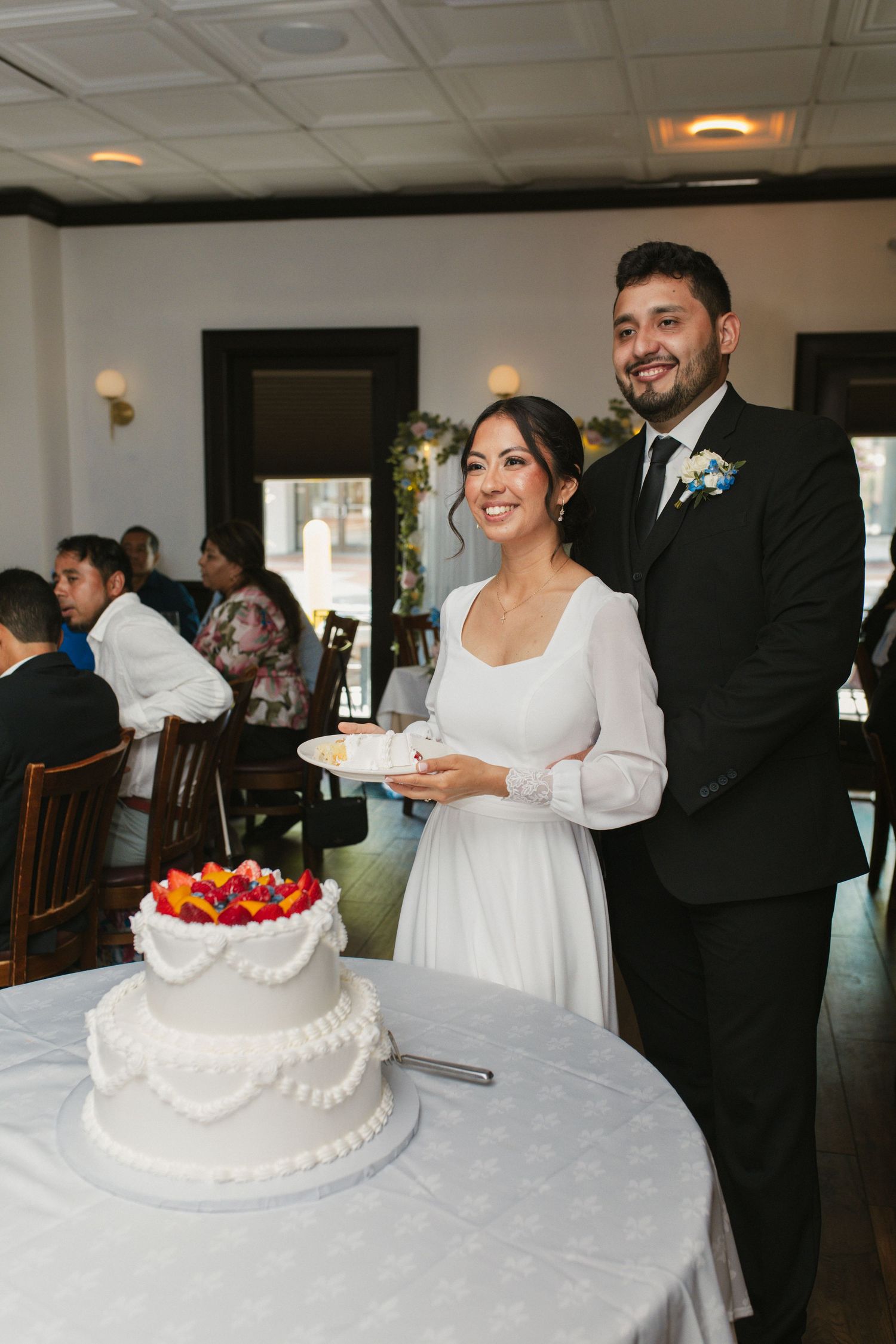 Smiling bride and groom pose together while cutting their two-tier white wedding cake at reception.