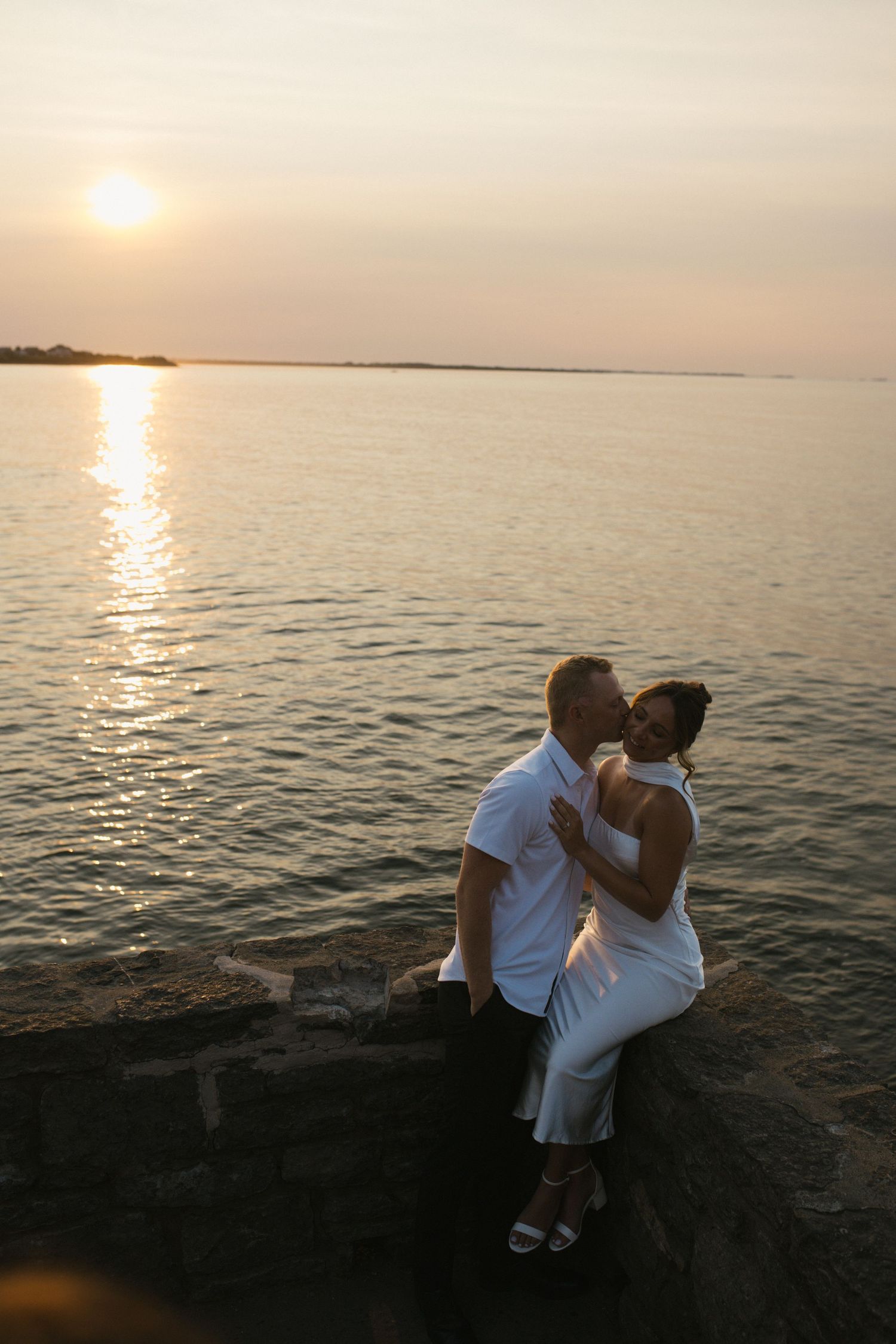 Couple sharing a romantic kiss at sunset by the water, dressed in white outfits on a rocky shoreline.