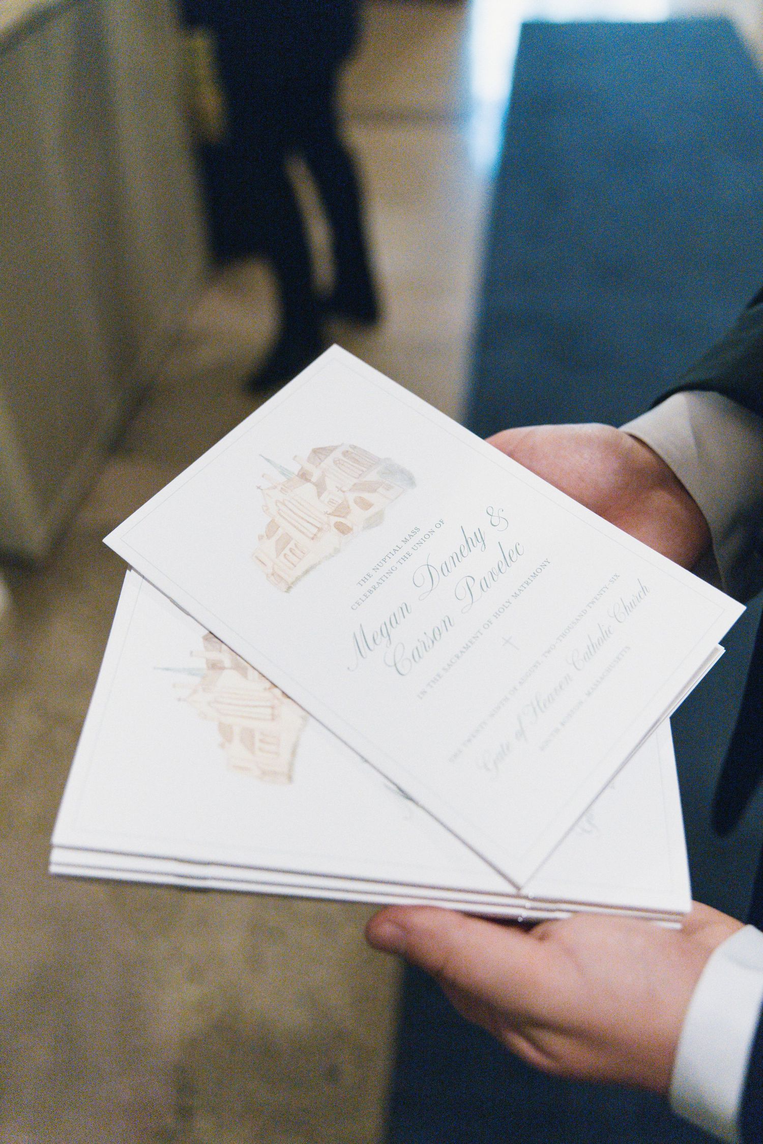Hands holding elegant white wedding ceremony programs with gold illustrated details and couple's names in script font.