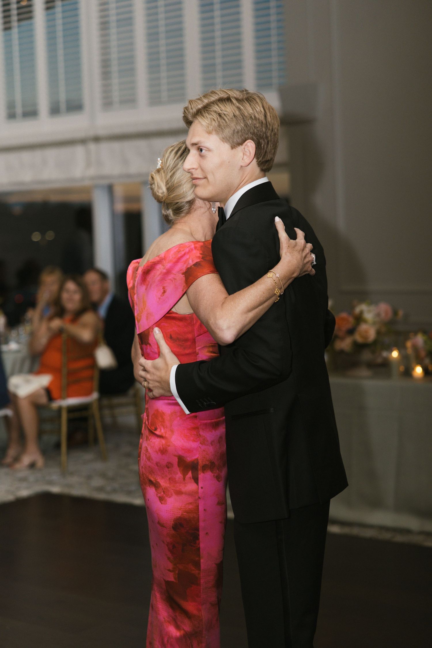 Groom in black tuxedo shares a mother-son dance with woman in vibrant pink floral gown.