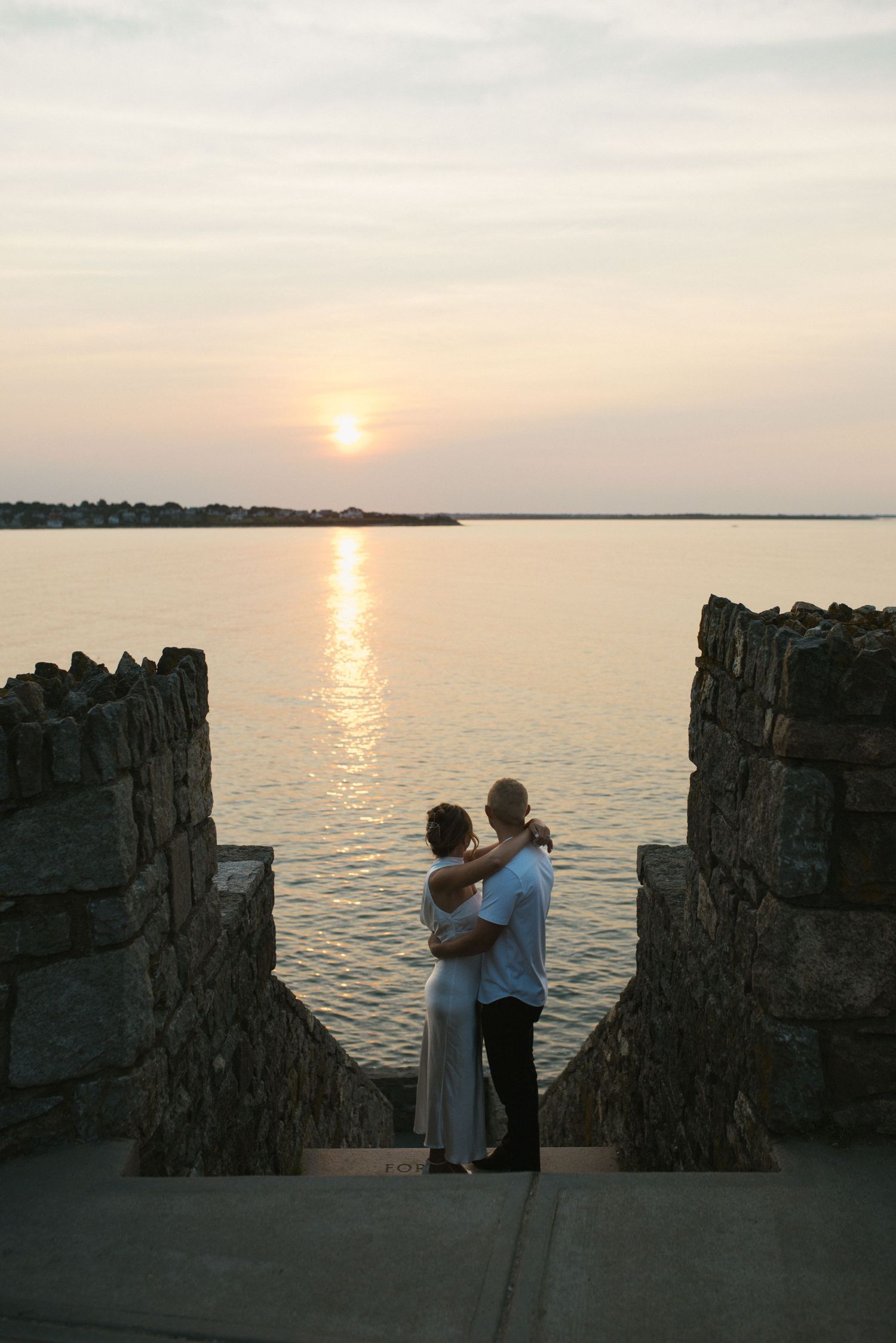 Couple embracing at sunset by stone walls overlooking calm ocean water, romantic waterfront scene at dusk.