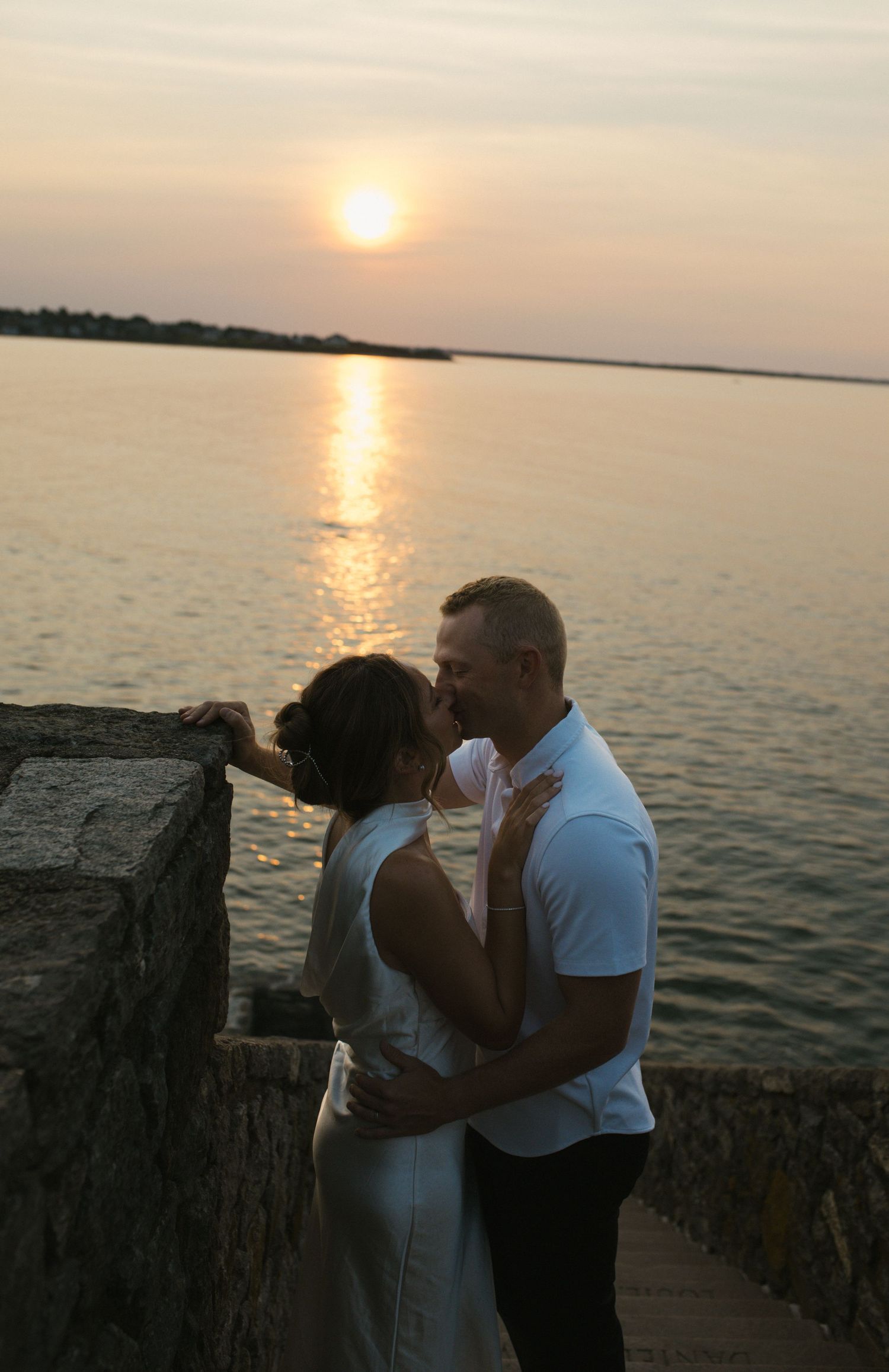 Couple sharing a romantic moment by stone wall at sunset over calm water, woman in white dress, man in white polo shirt.