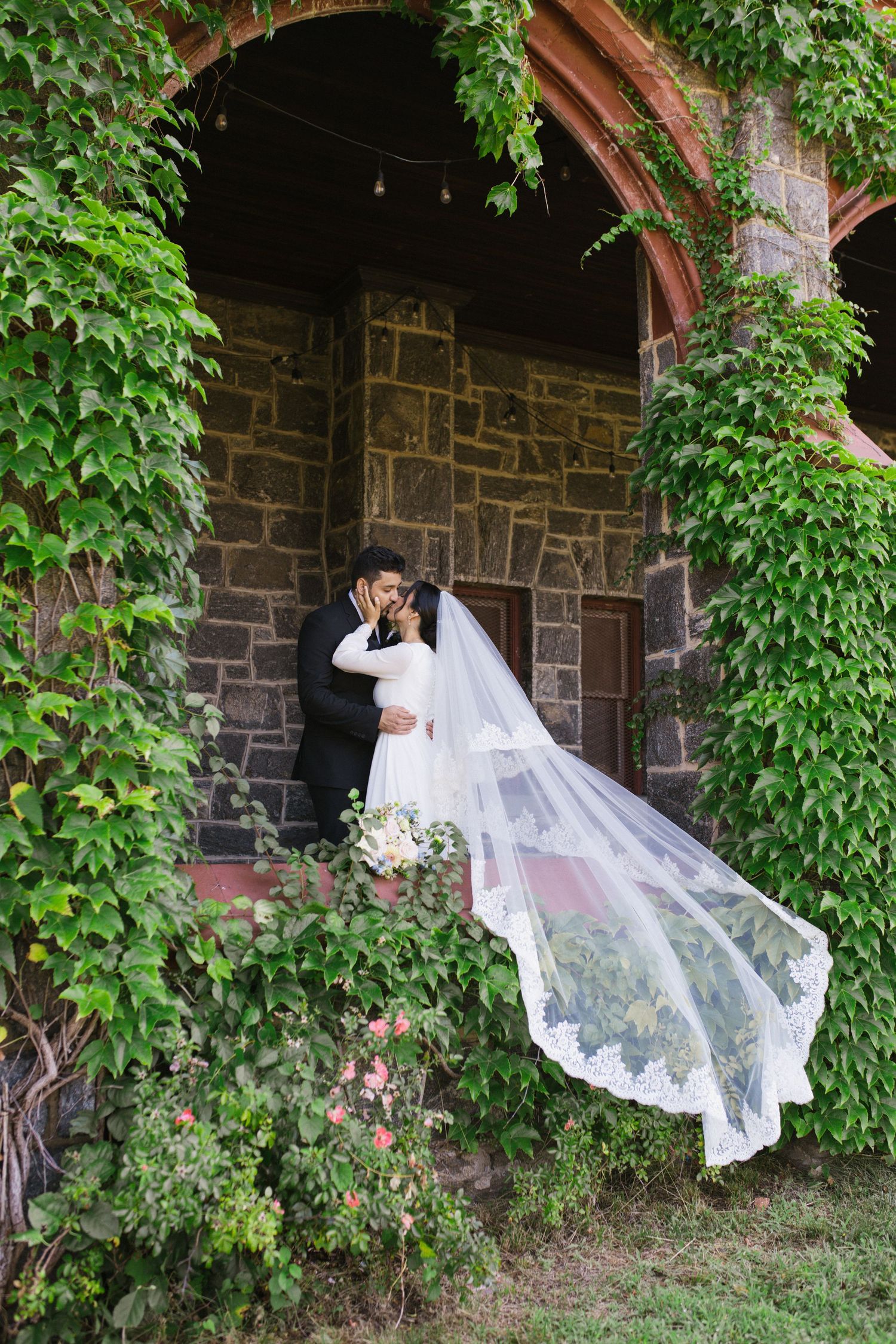 Bride and groom sharing a kiss in a romantic ivy-covered stone building, with the bride's lace veil flowing in the wind.