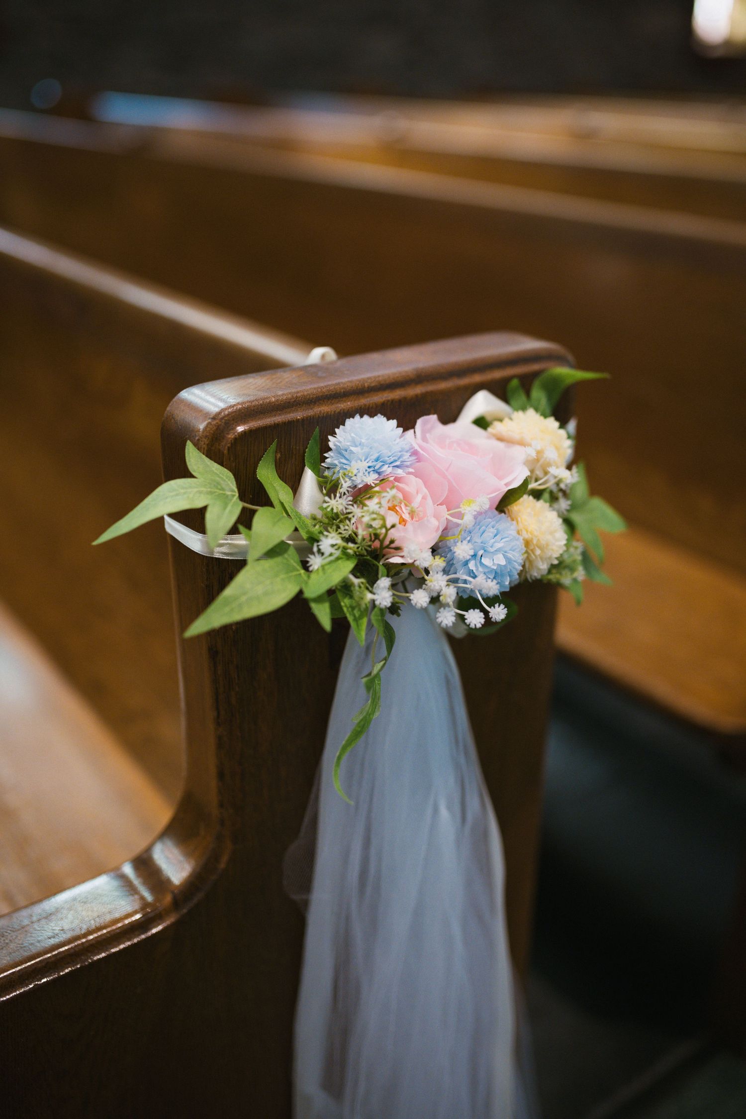 Floral pew decoration with pink, blue, and white flowers tied with tulle ribbon on wooden church bench.