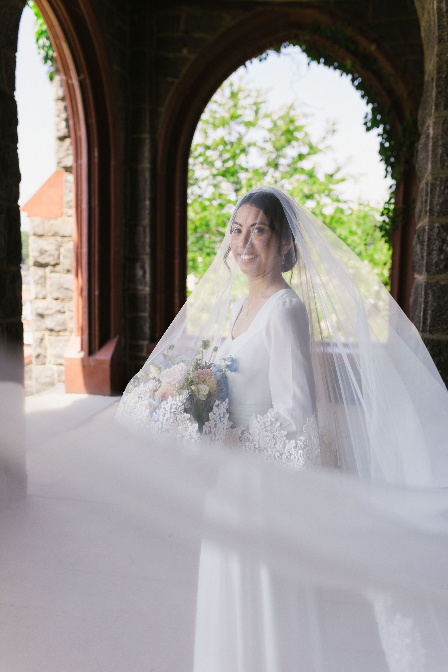 Smiling bride in white gown and veil holds bouquet under stone archway on wedding day.