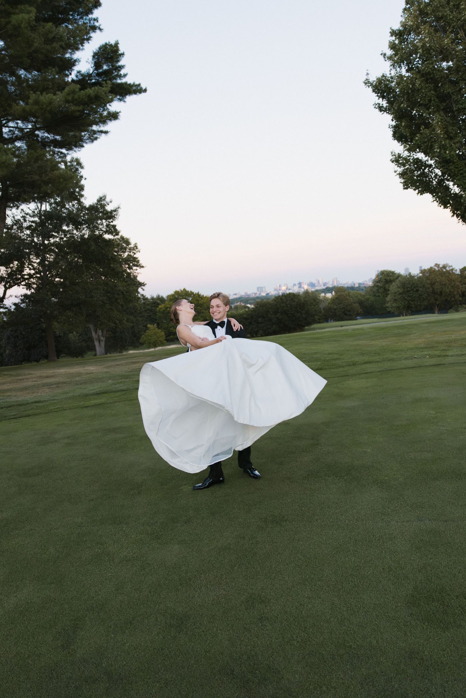 Groom twirls bride in flowing white wedding dress on a green golf course at dusk.