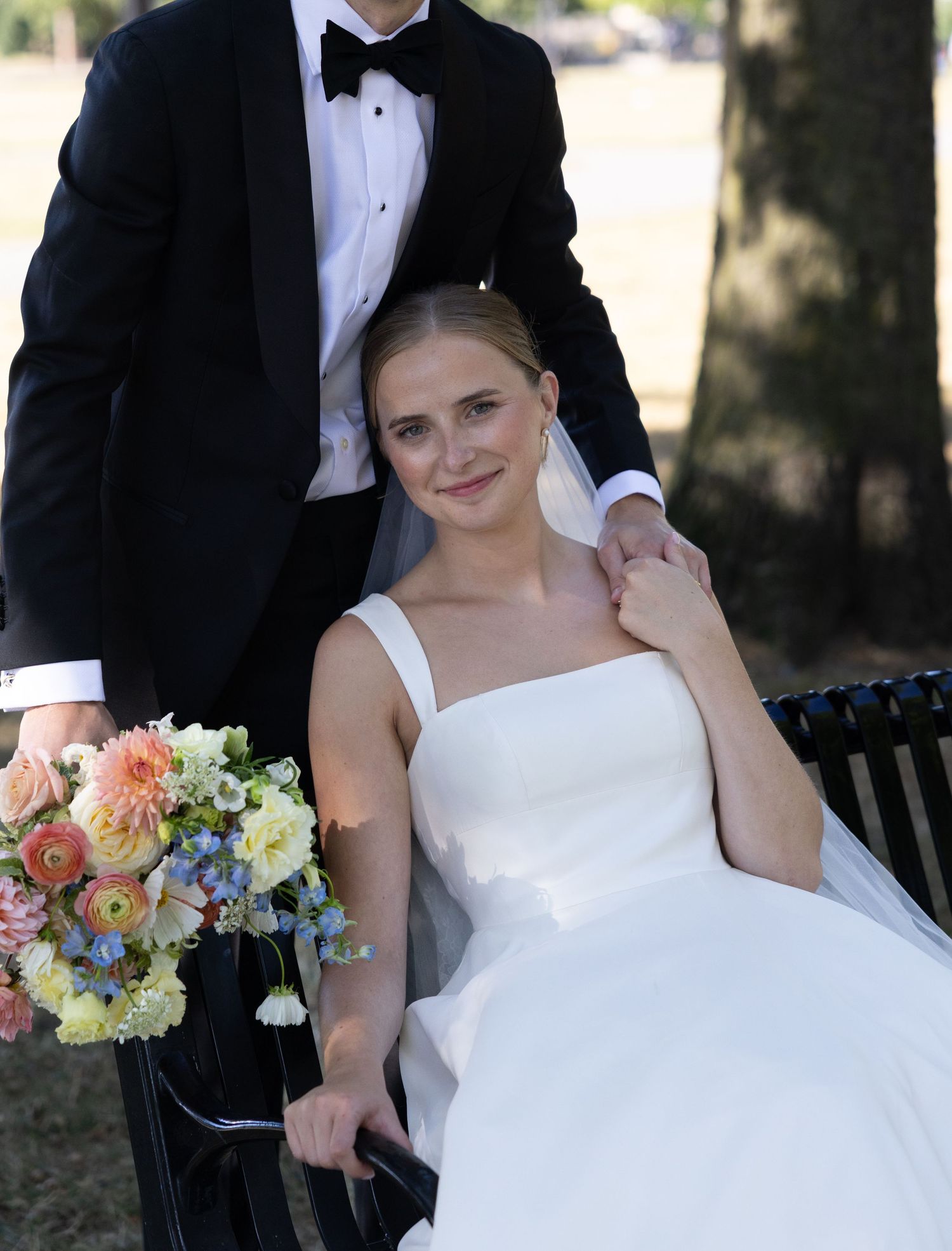 Bride in white gown holds colorful bouquet while groom in black tuxedo stands behind her on their wedding day.