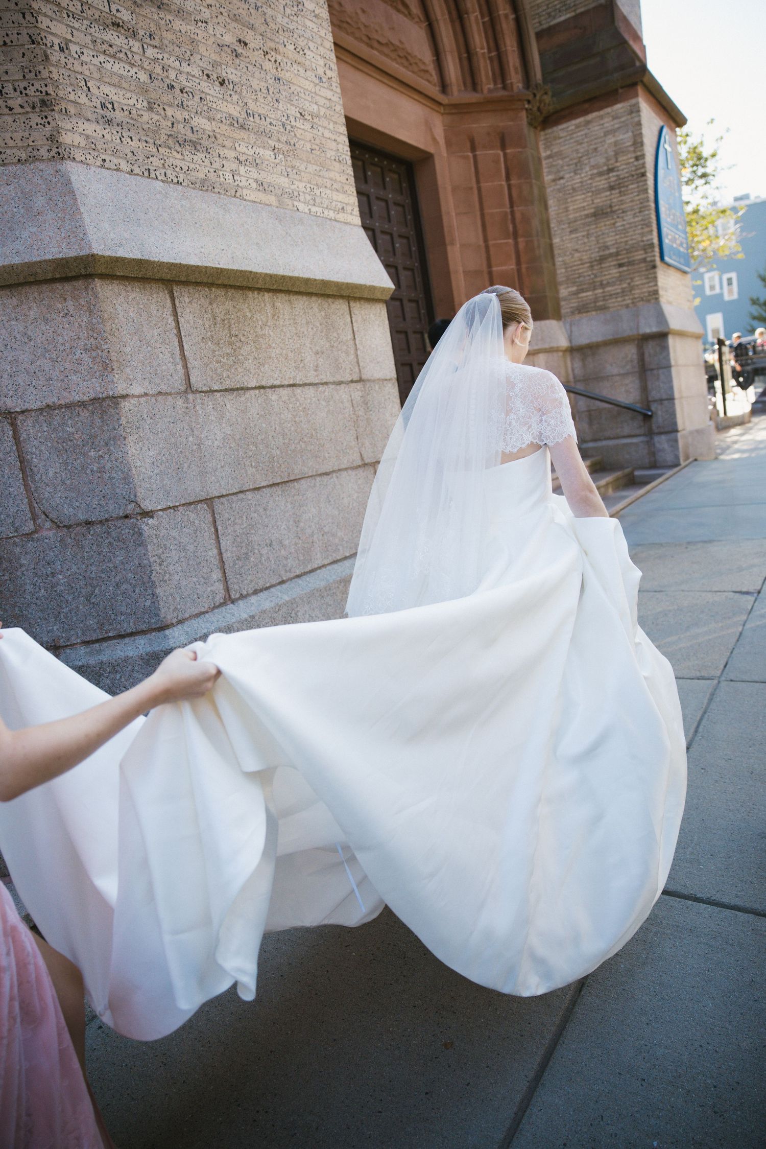 Bridesmaid holding wedding dress train as bride walks along sidewalk outside stone church.