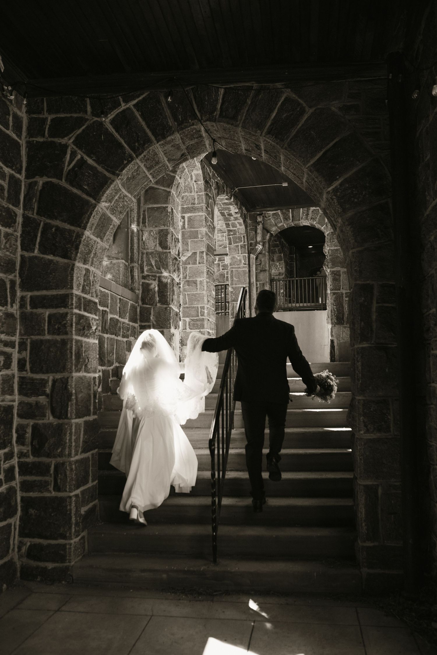 Bride and groom silhouetted walking through stone arch tunnel stairway in black and white.