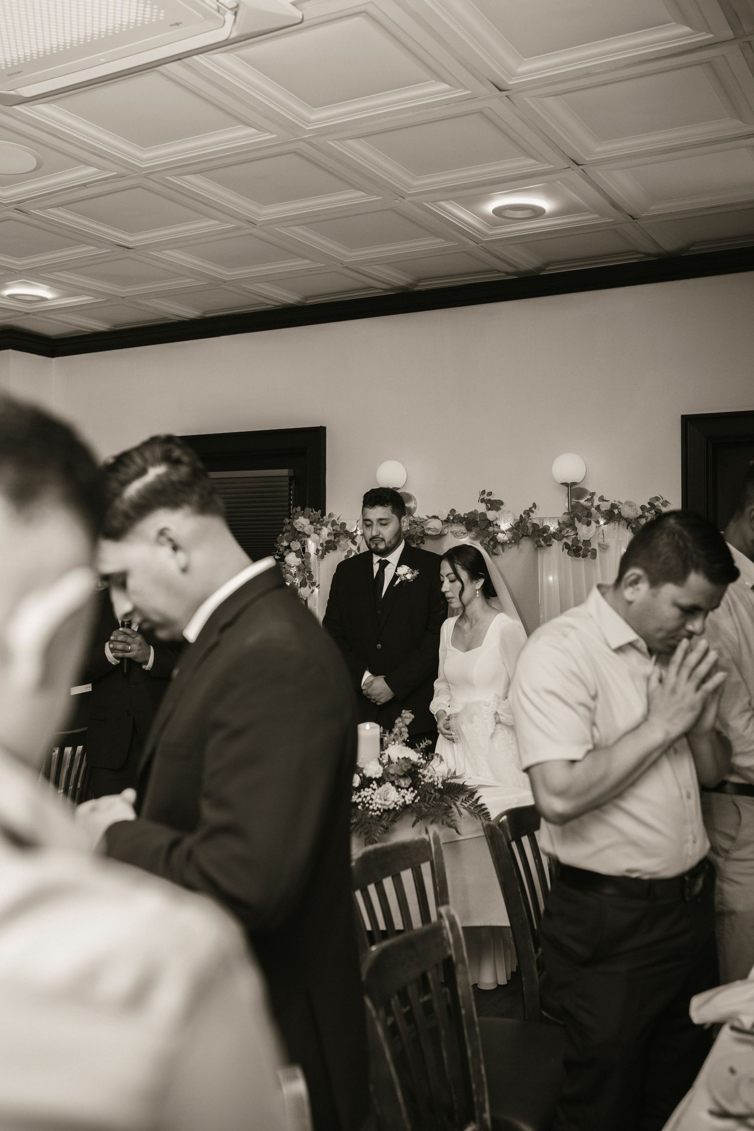 Black and white photo of bride and groom standing at altar while guests pray at reception.
