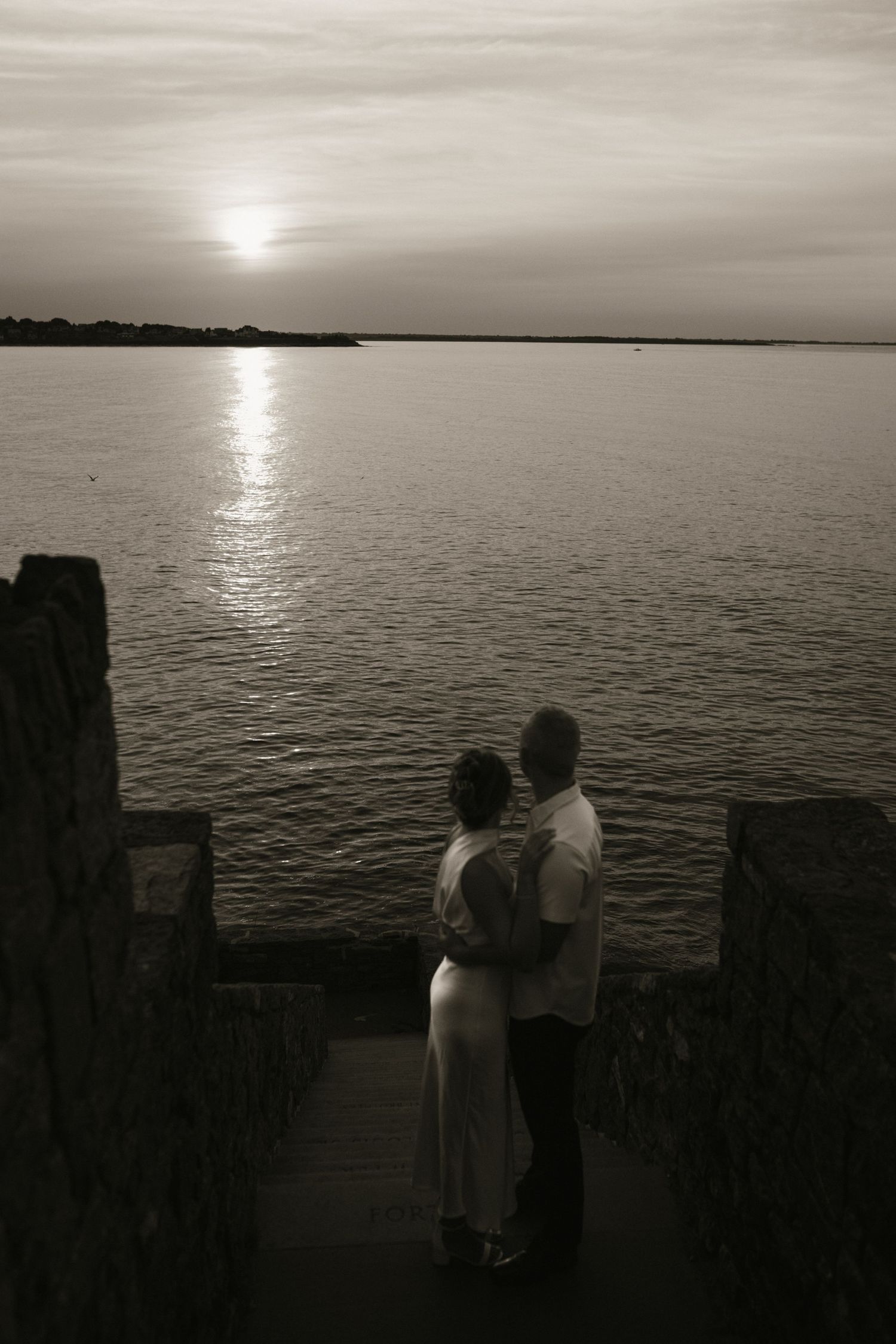 Couple embracing at sunset overlooking calm ocean waters from stone wall overlook, black and white romantic photo.