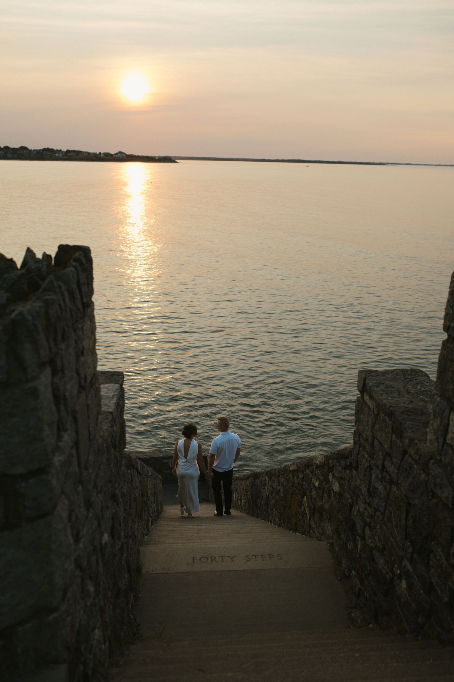 Couple watching golden sunset over calm ocean waters from stone stairway leading to the sea.
