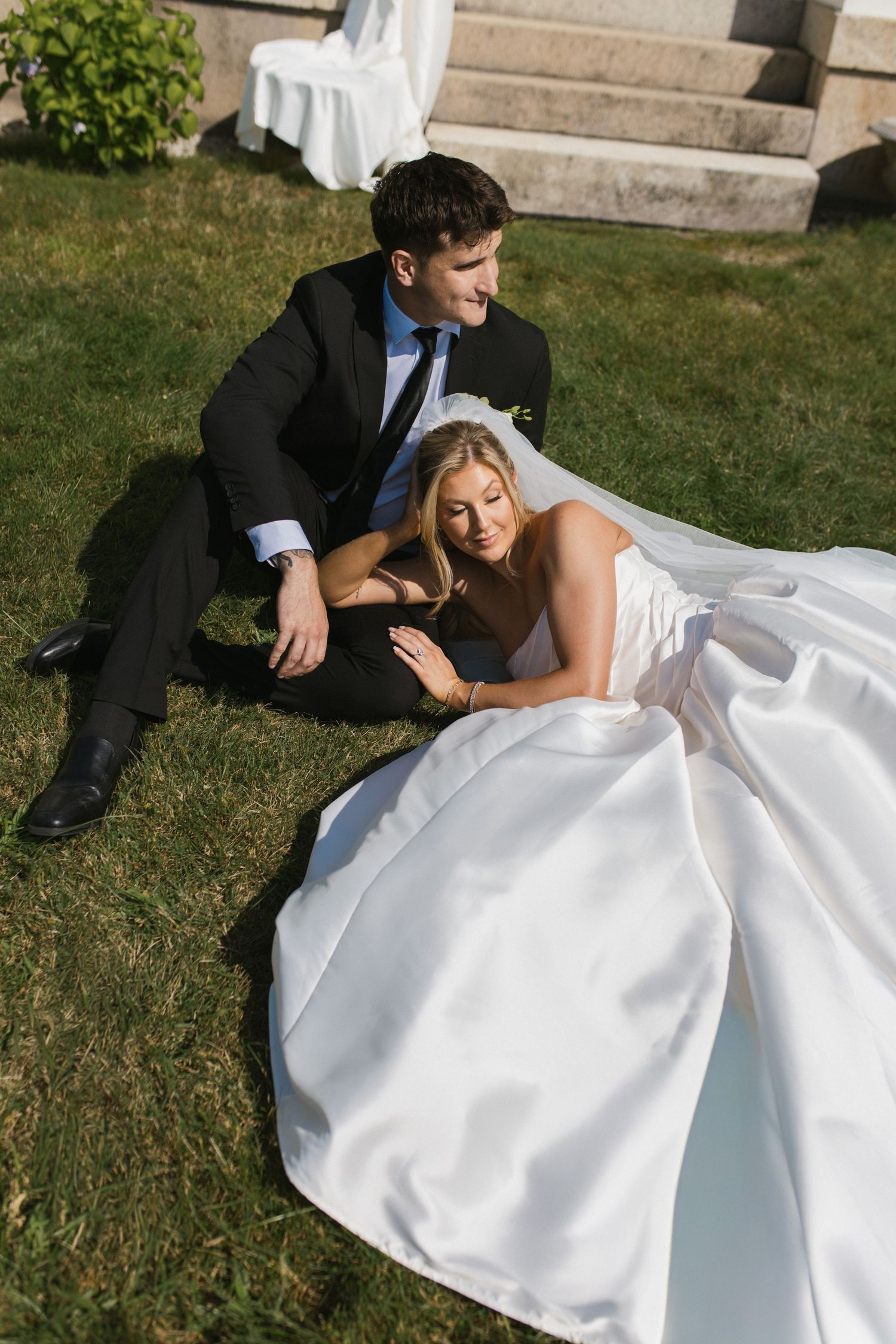Bride in a voluminous white satin gown sits with groom in black suit on green grass beside stone steps.