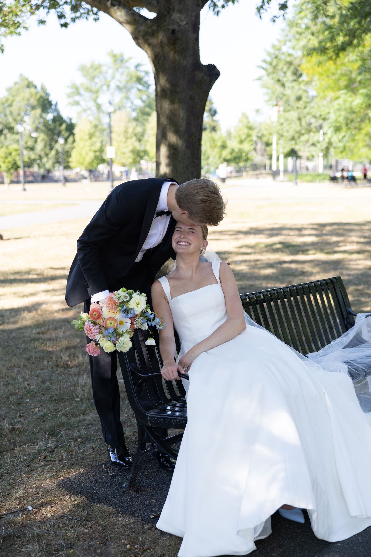 Groom in black tuxedo kissing bride on cheek as she sits on park bench holding colorful bouquet.