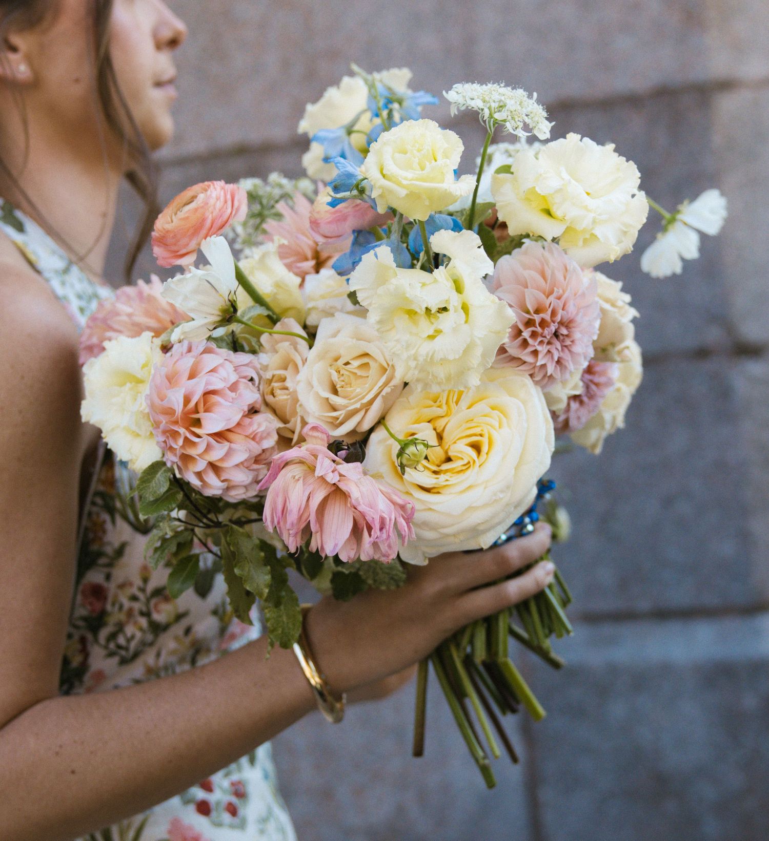 Woman holding a lush bridal bouquet of pastel pink dahlias, cream roses, and blue flowers in soft natural light.