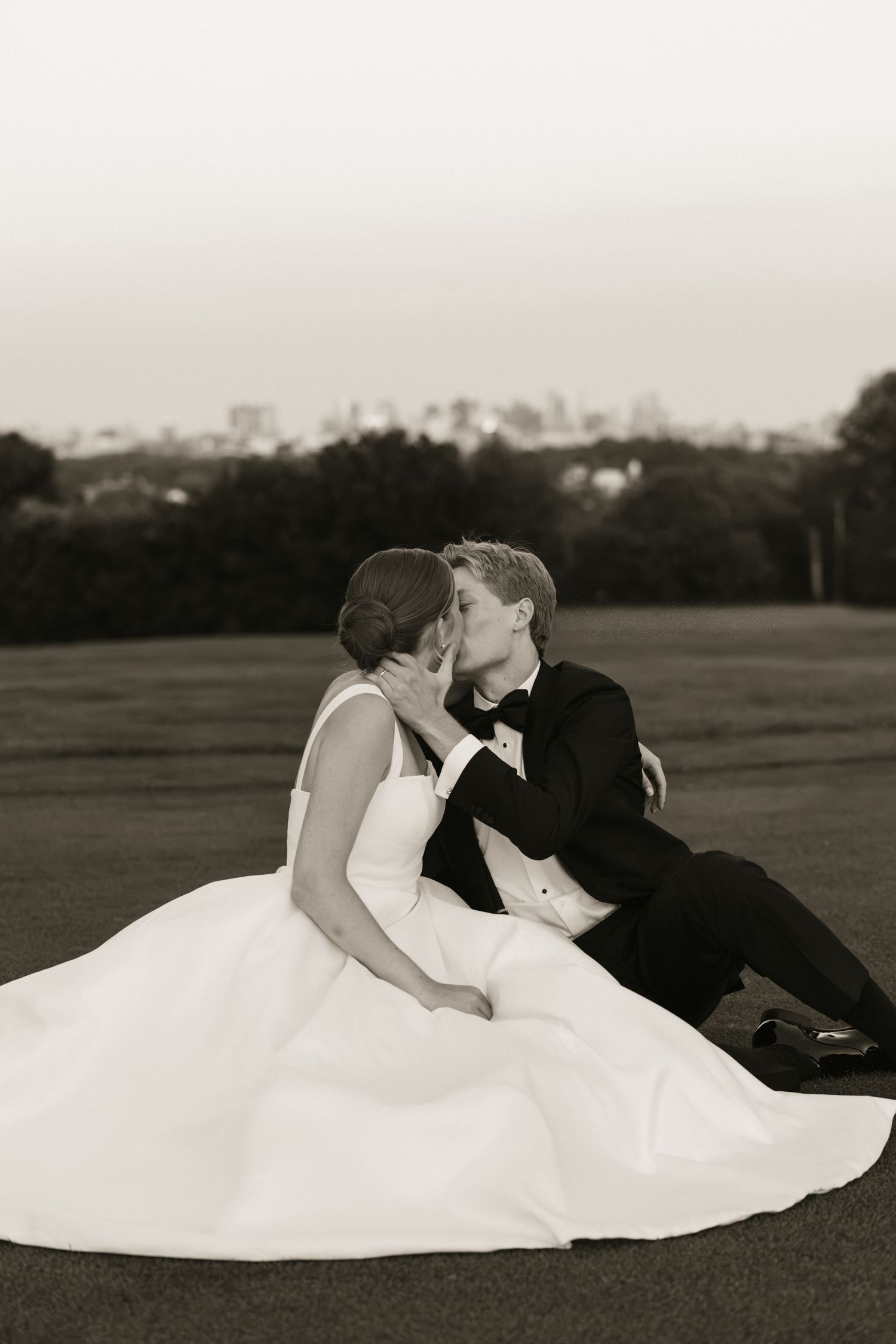 Black and white photo of bride and groom sharing a kiss while seated on a golf course with city skyline backdrop.