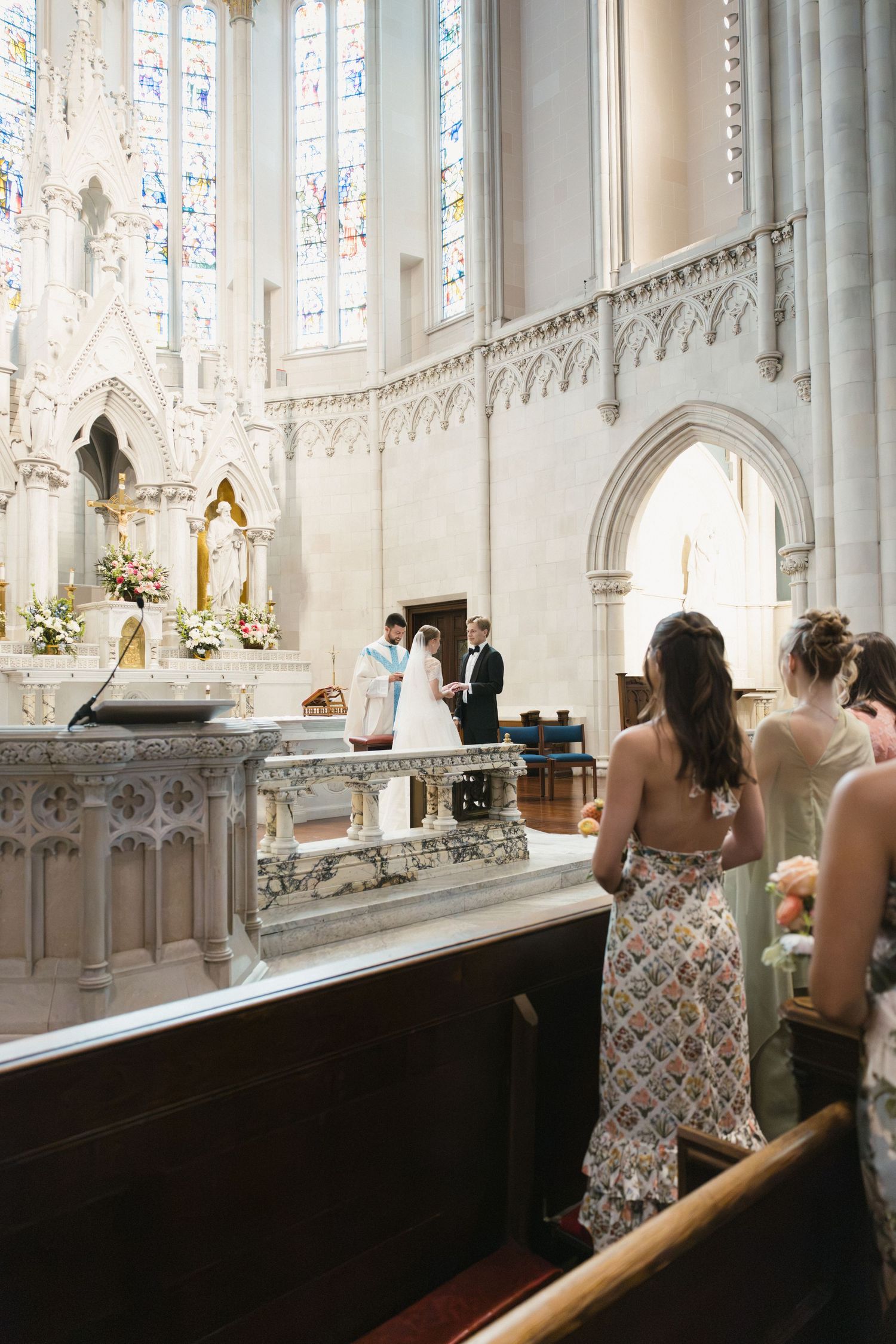 Wedding ceremony at ornate white Gothic church altar with bride, groom, and bridesmaids in patterned dresses.