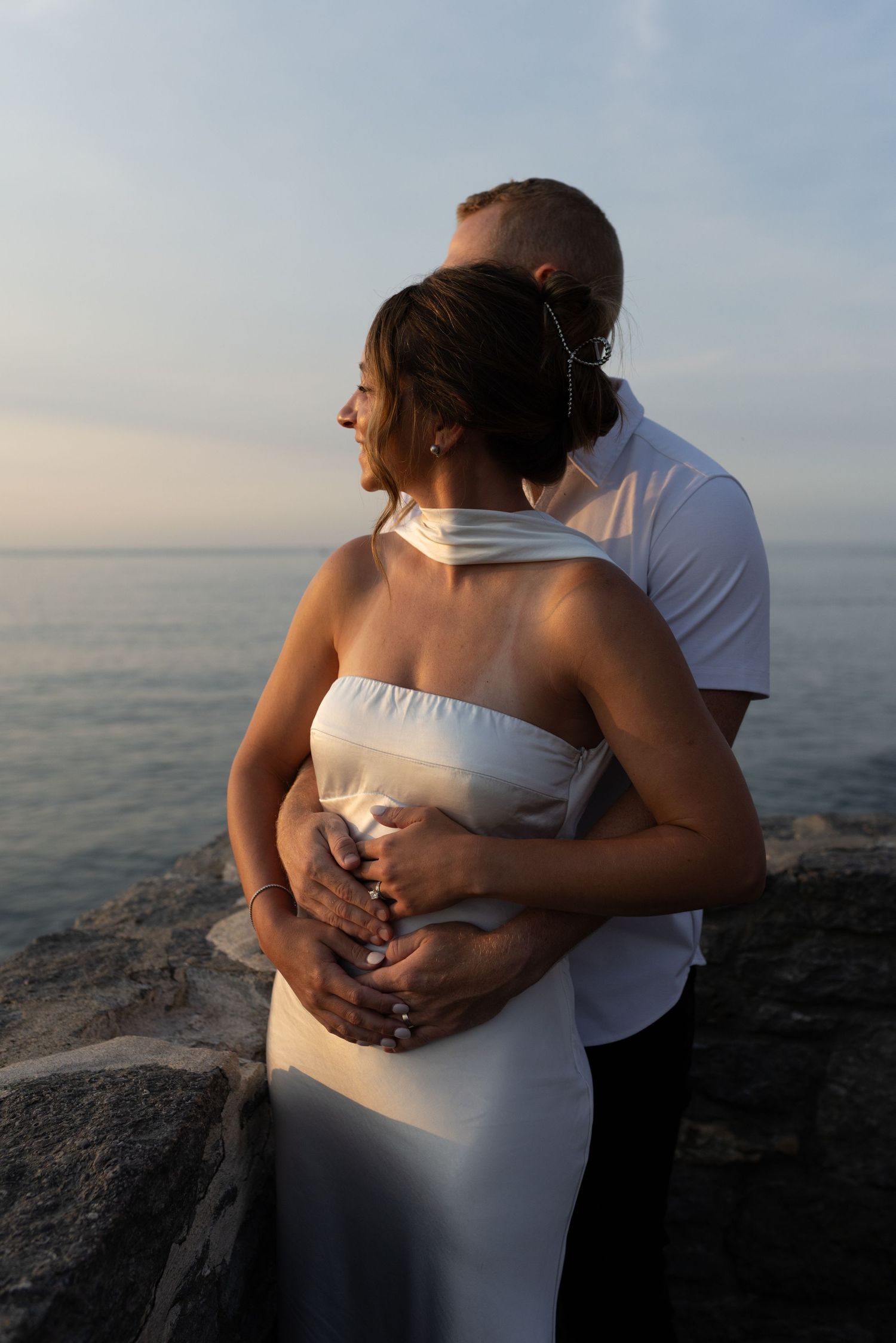Man tenderly embracing woman in white halter dress on rocky shoreline at dusk.