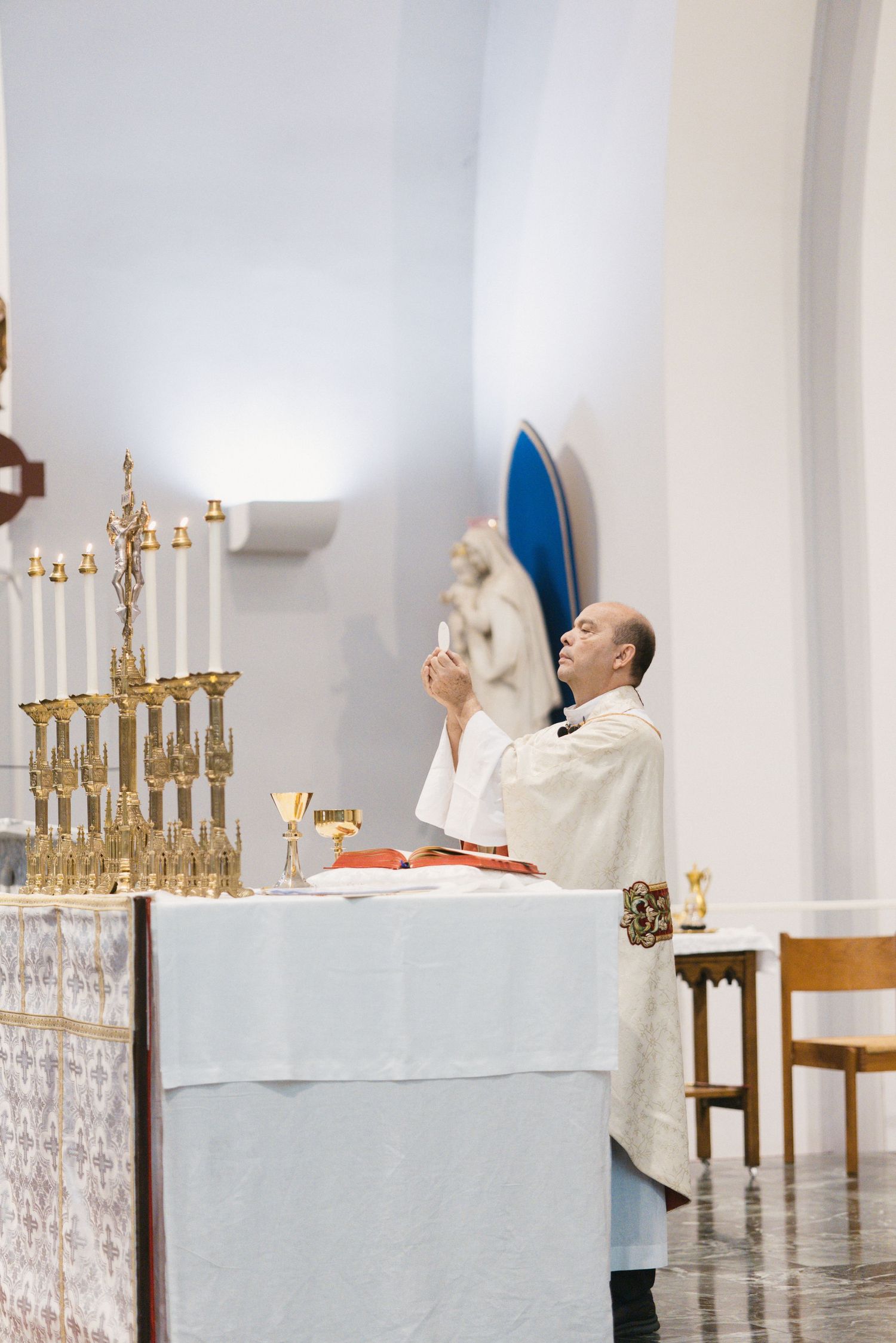 Catholic priest in white vestments prays at altar with gold candelabras during Mass.