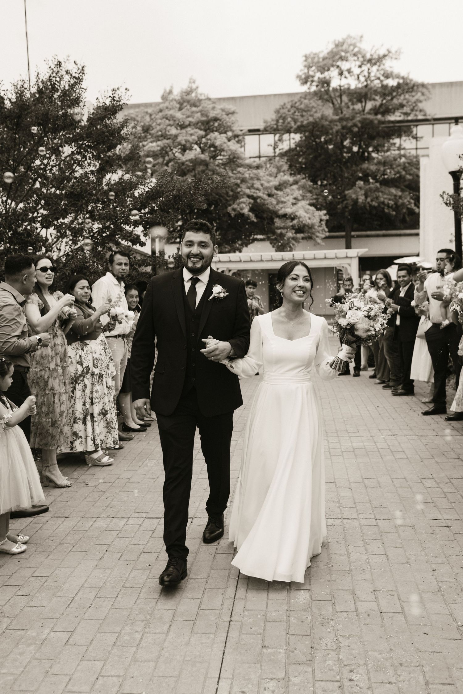 Newlyweds walk hand-in-hand through cheering guests outside after wedding ceremony, black and white photo.
