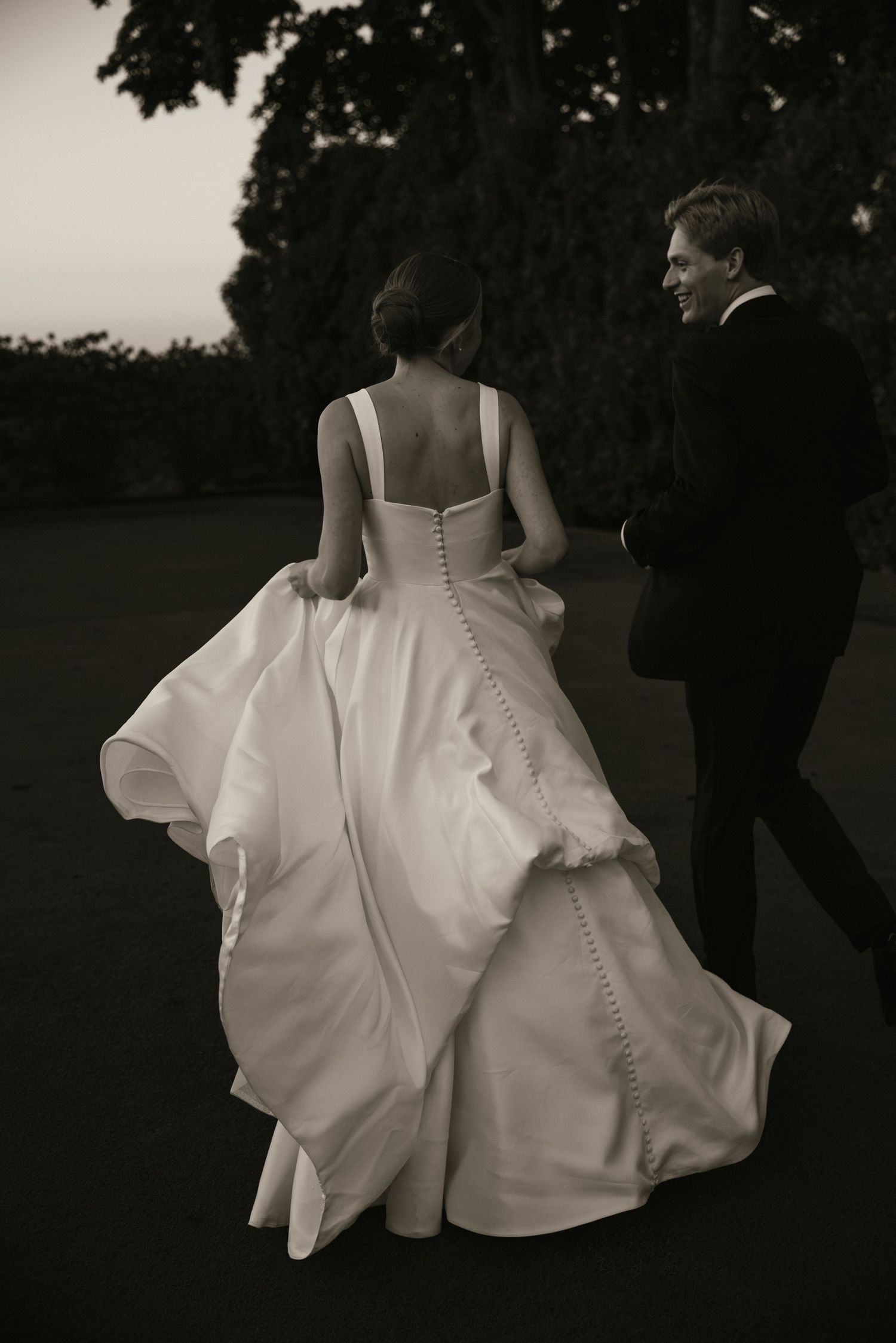Bride in a button-back white gown walks with groom at dusk in an elegant black and white photo.