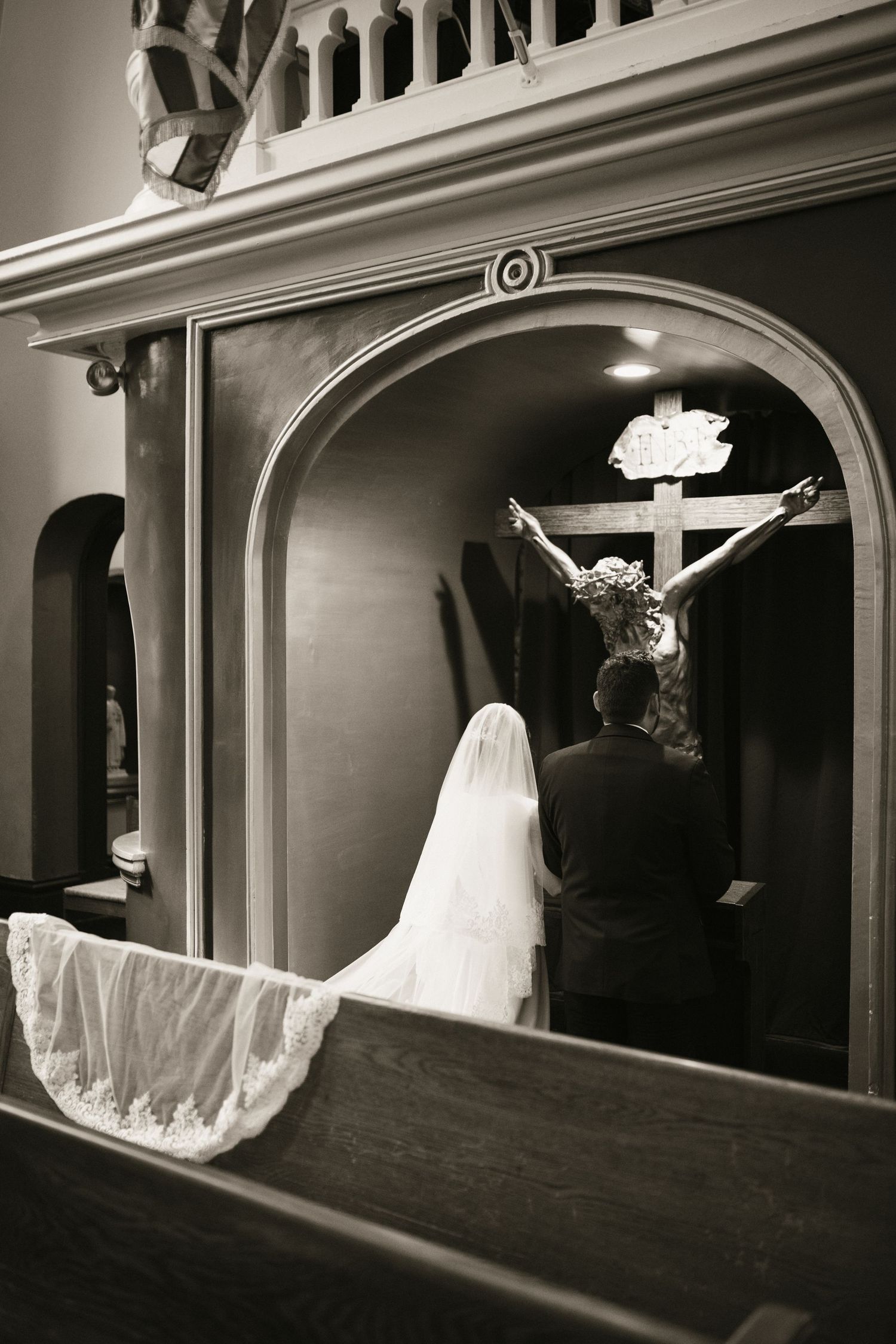 Black and white photo of bride and groom kneeling in prayer before crucifix in church confessional.