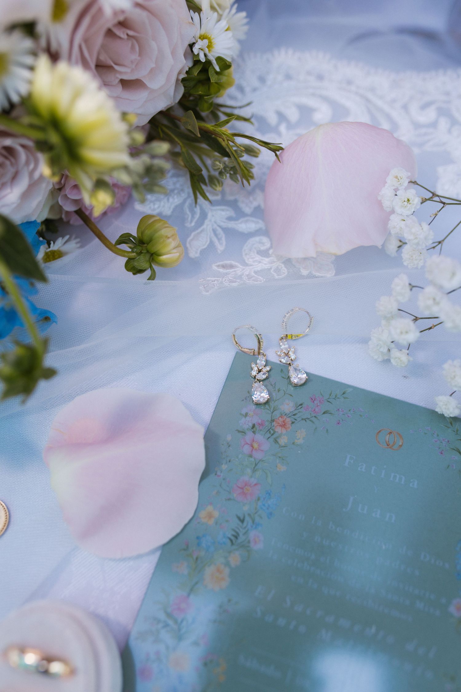 Wedding flat lay with teal menu card, crystal earrings, pink rose petals and soft floral bouquet.
