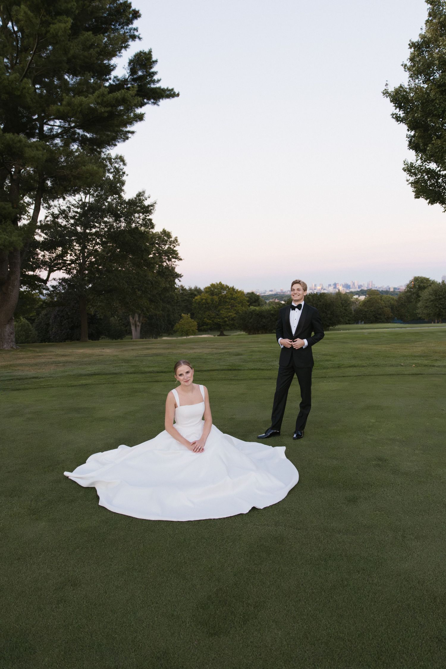 Bride in flowing white ballgown sits on green lawn while groom stands in black tuxedo at dusk.