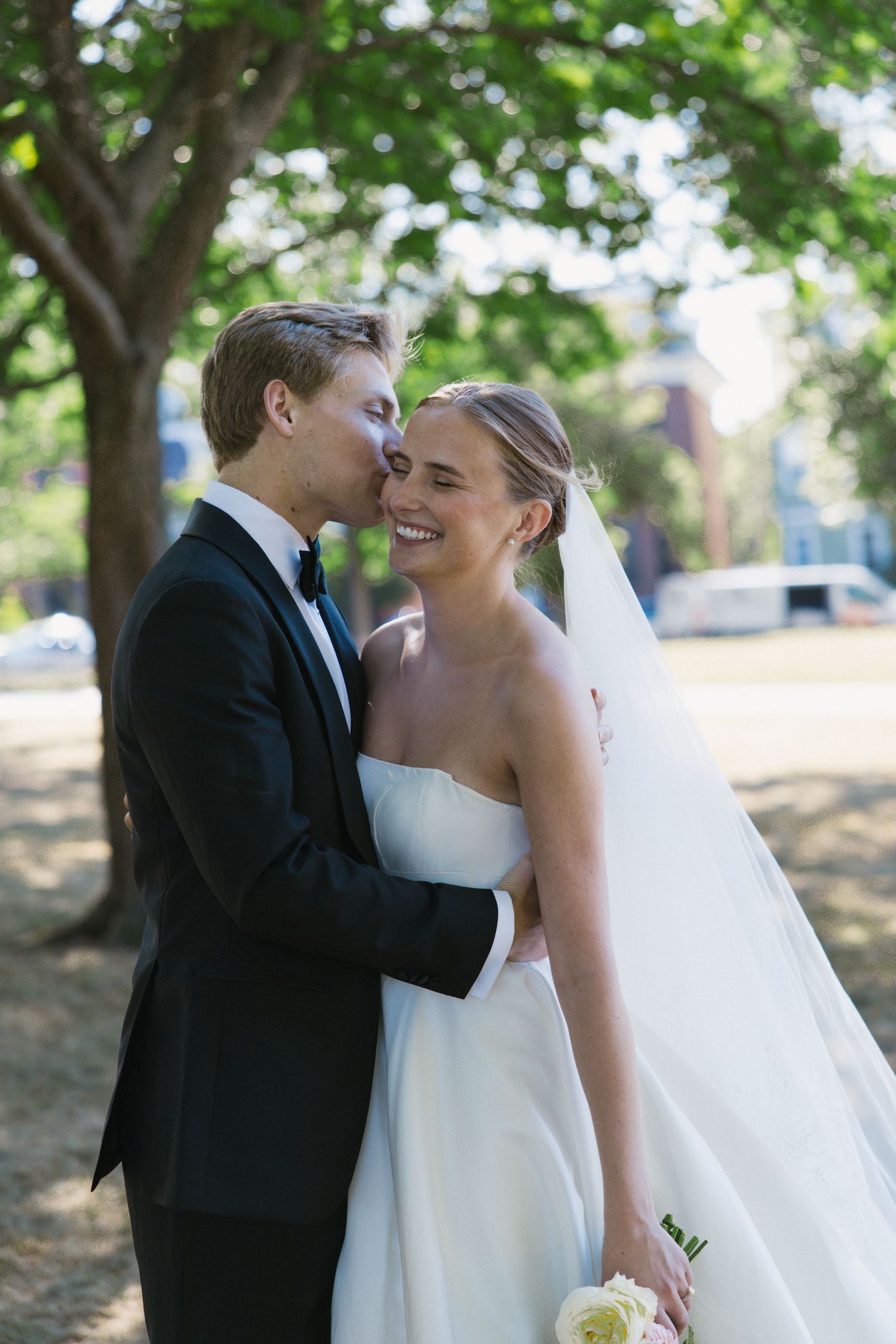 Happy bride in strapless white gown laughs as groom in black tuxedo kisses her cheek outdoors.