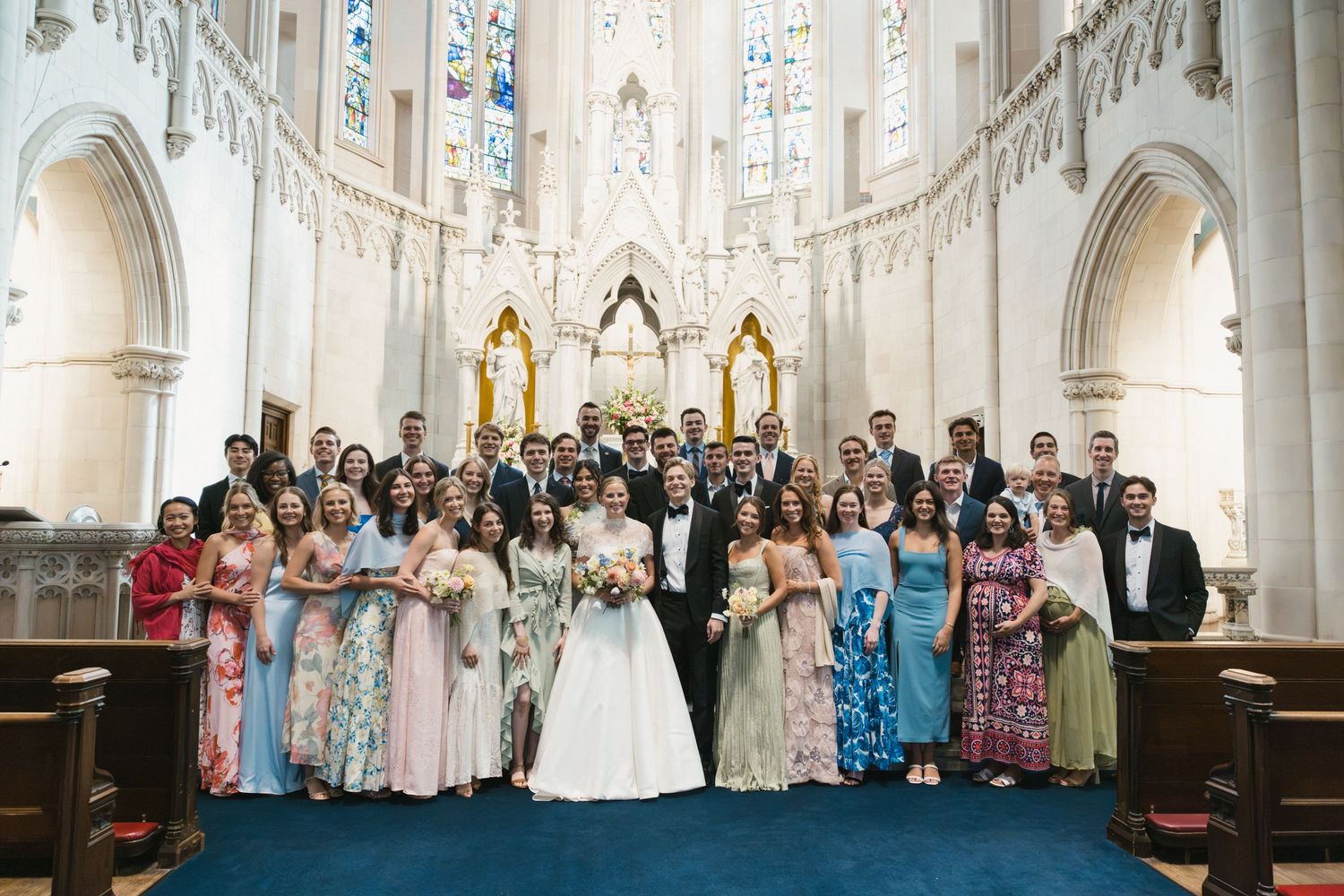 Wedding party poses together in an ornate church with blue carpet, stained glass windows, and golden altar decorations.