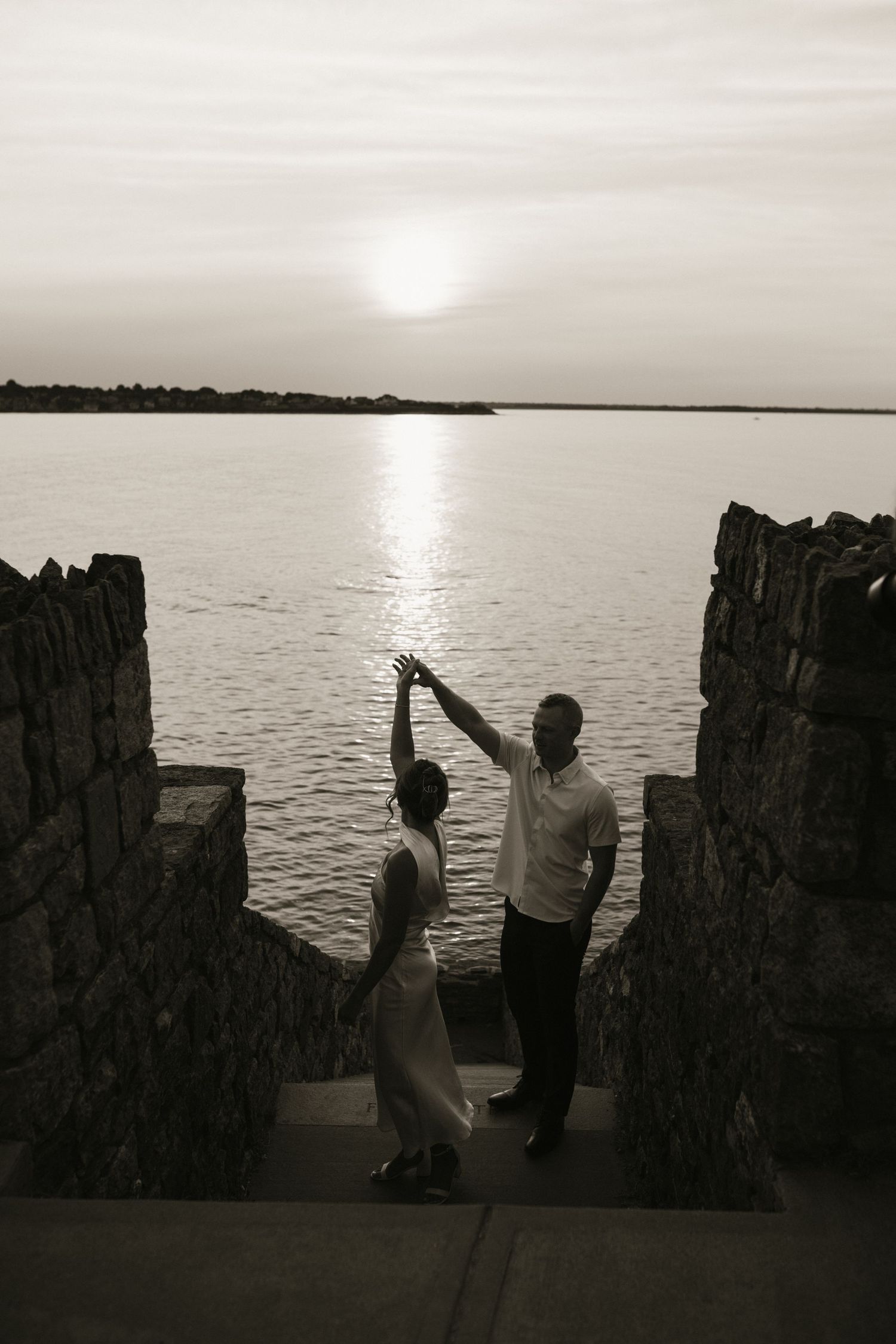 Black and white photo of couple dancing at sunset by stone ruins near tranquil ocean.