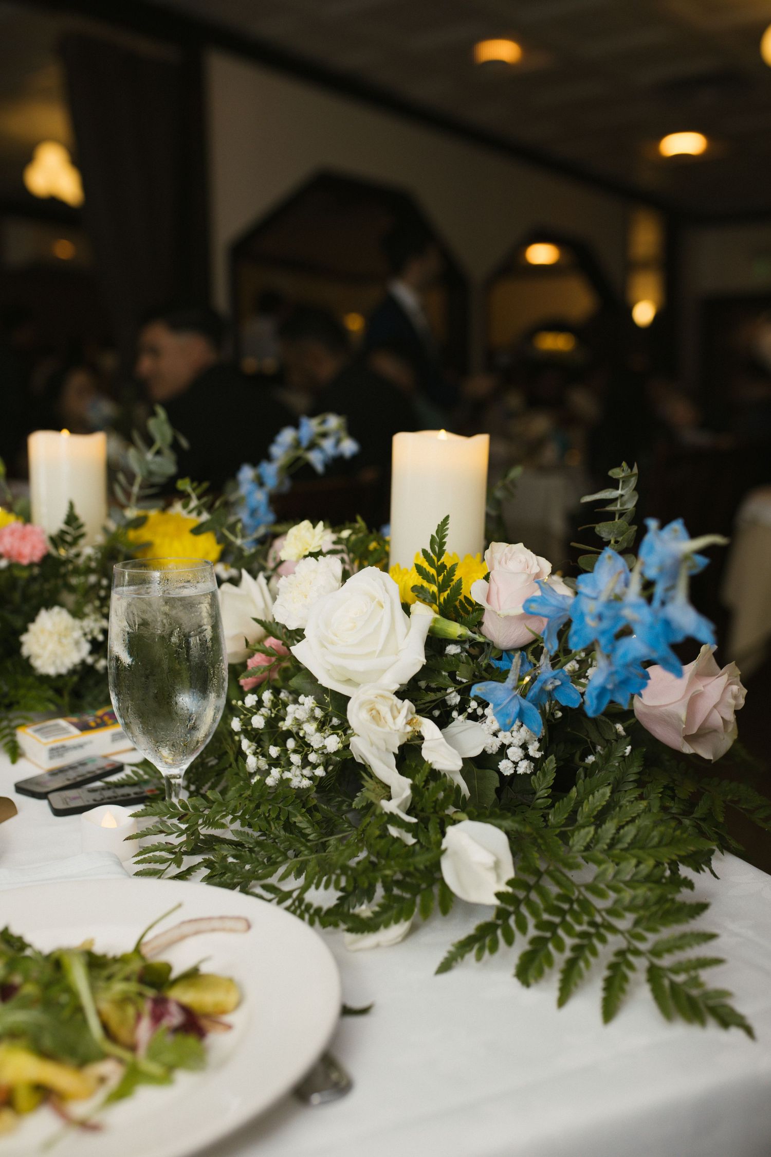 Elegant wedding reception centerpiece with white candles, roses, blue flowers, and lush greenery.
