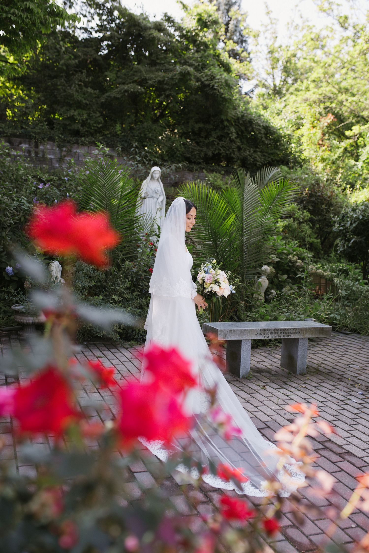 Elegant bride in white gown and cathedral veil walks through lush garden with bouquet near statue.
