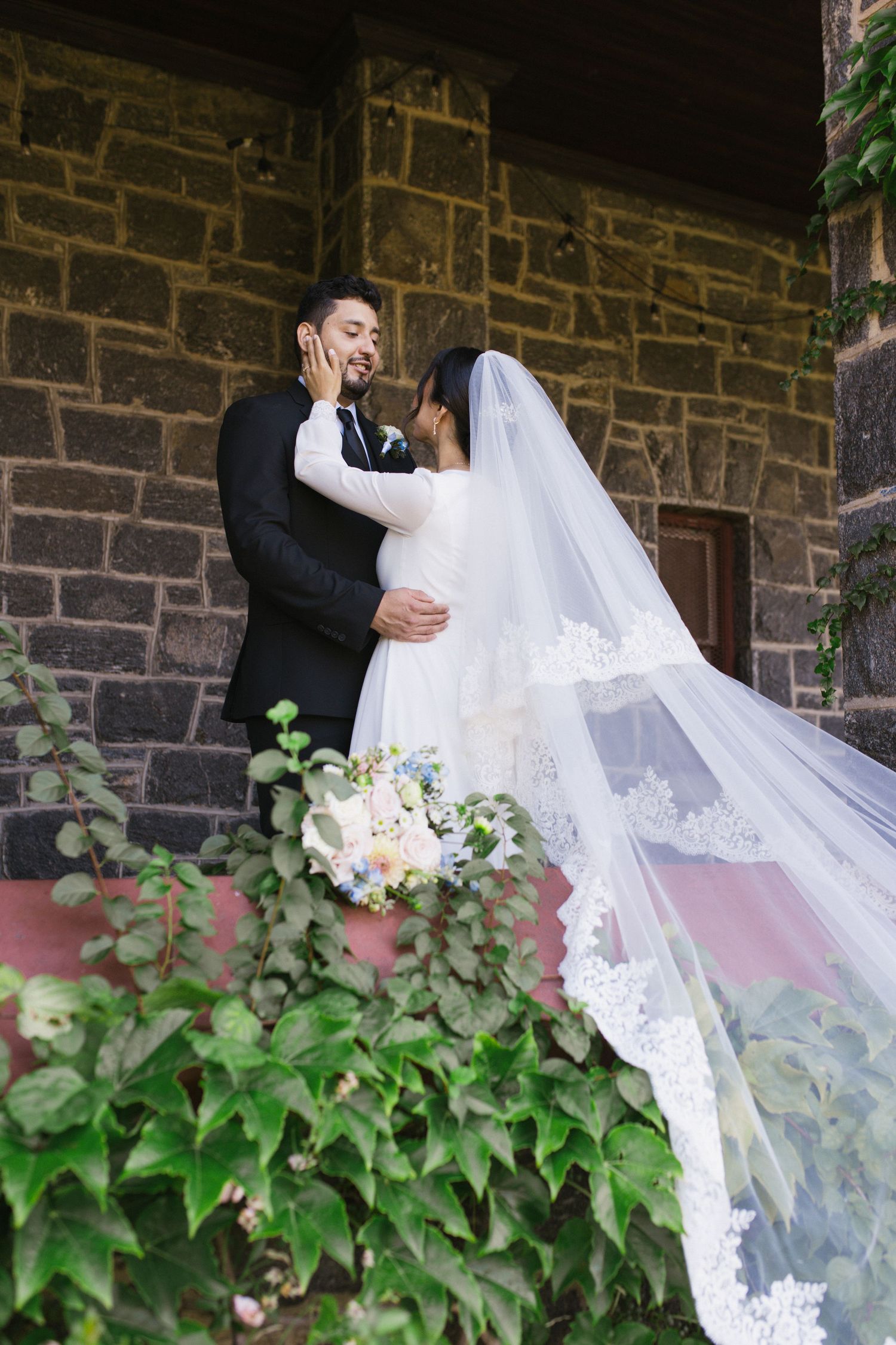 Bride and groom share a romantic embrace on stone church steps, her lace veil flowing gracefully.