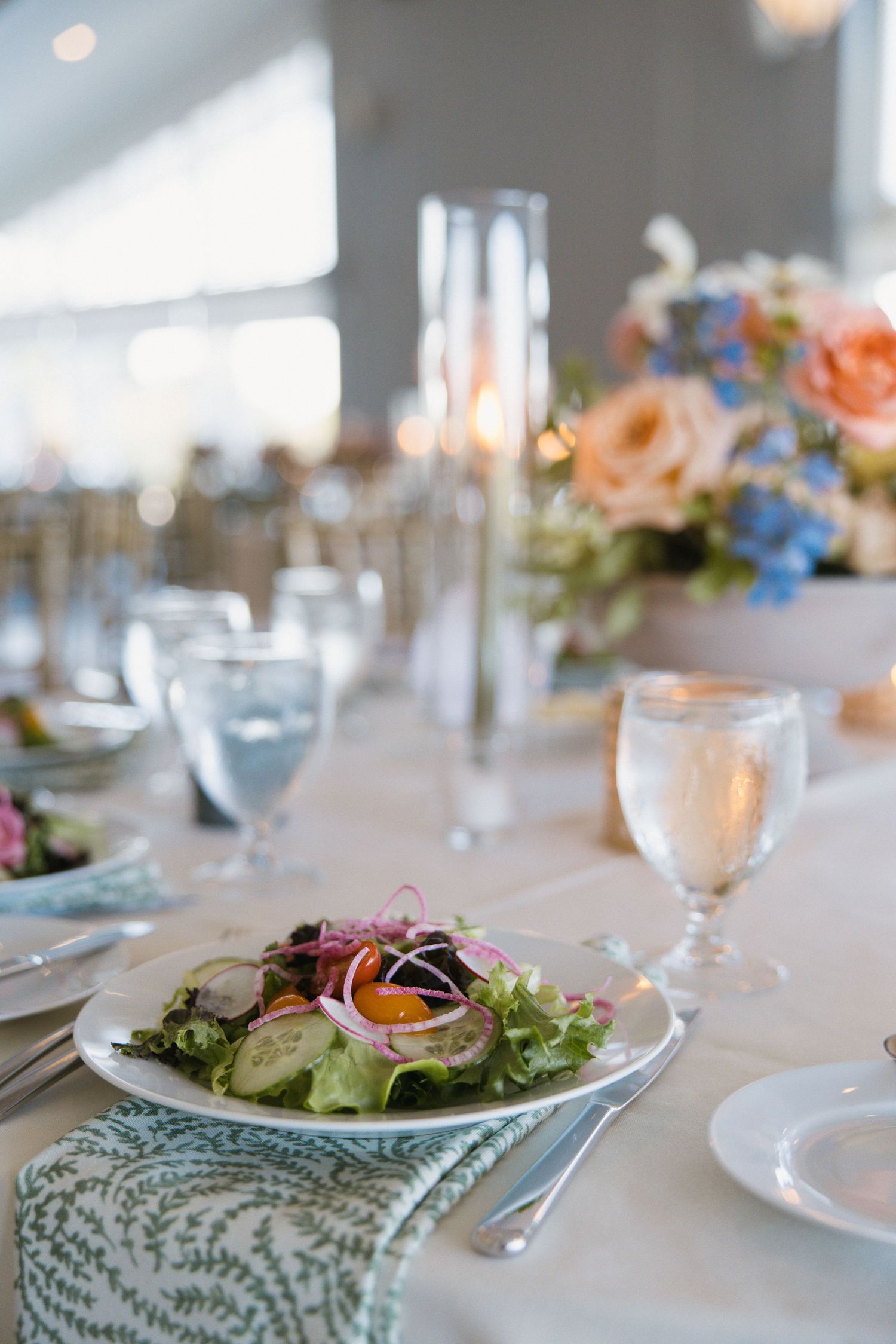 Elegant wedding reception table with fresh salad, floral centerpieces in peach and blue, and glassware.