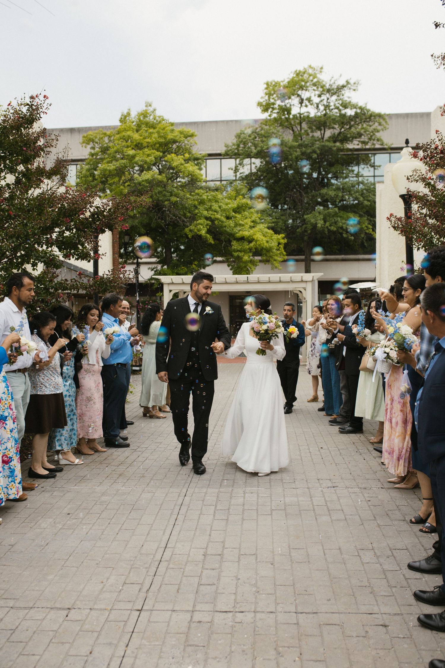 Newlyweds walk through bubble sendoff surrounded by cheering guests outside after wedding ceremony.
