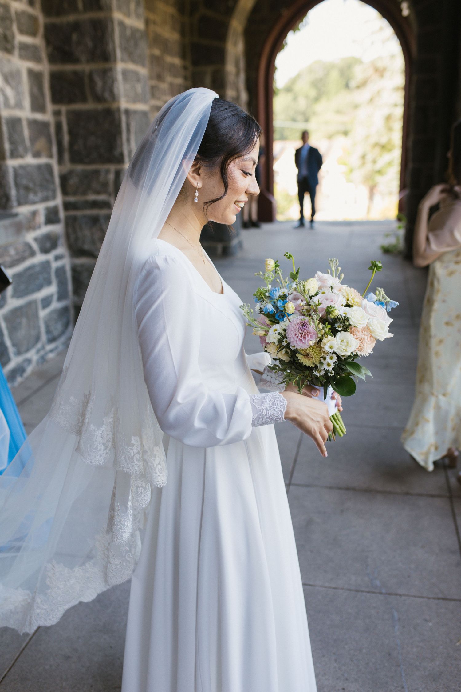 Elegant bride in long-sleeve white gown holds colorful bouquet in stone archway.