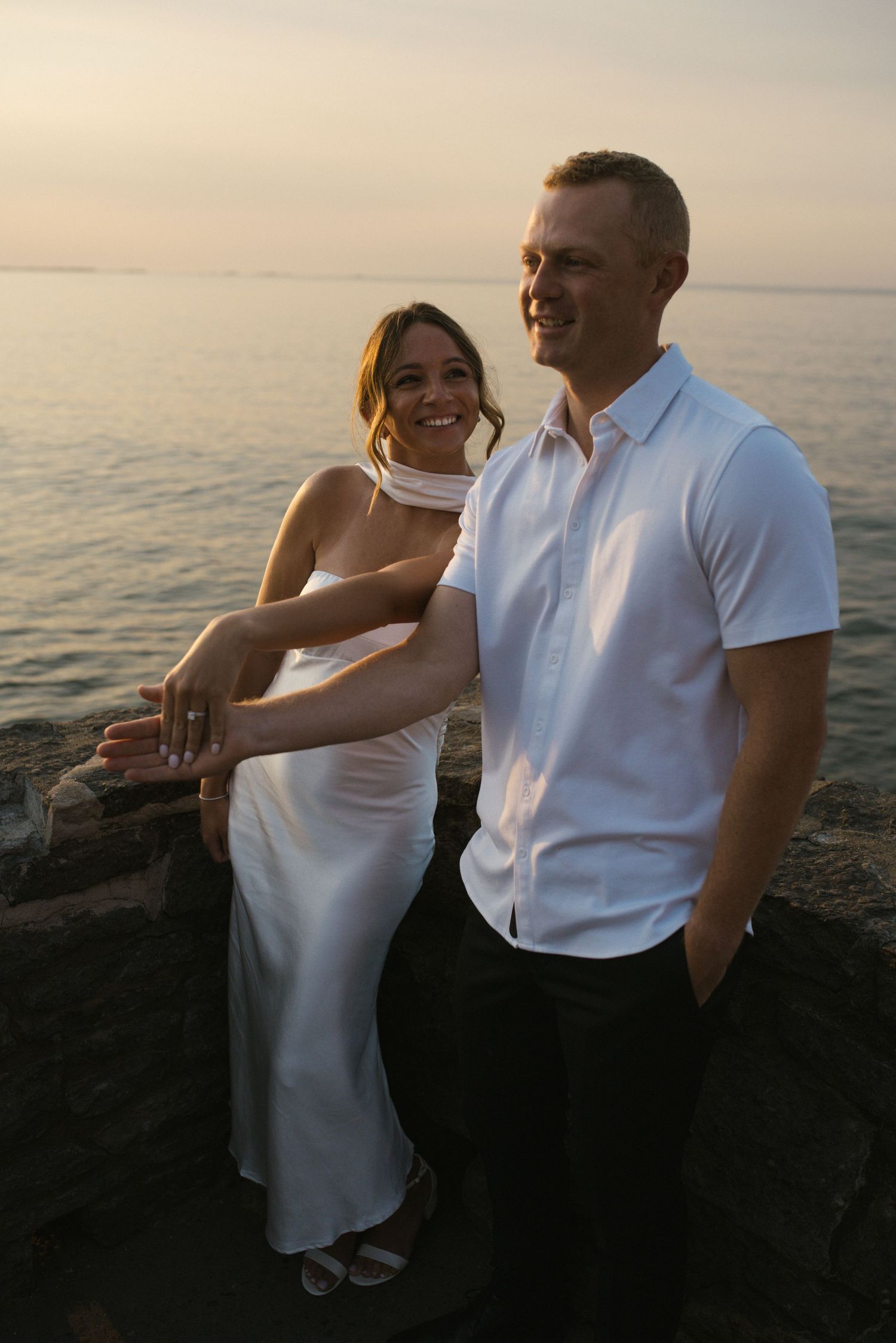 Smiling engaged couple posing on rocky waterfront at sunset, woman showing ring.
