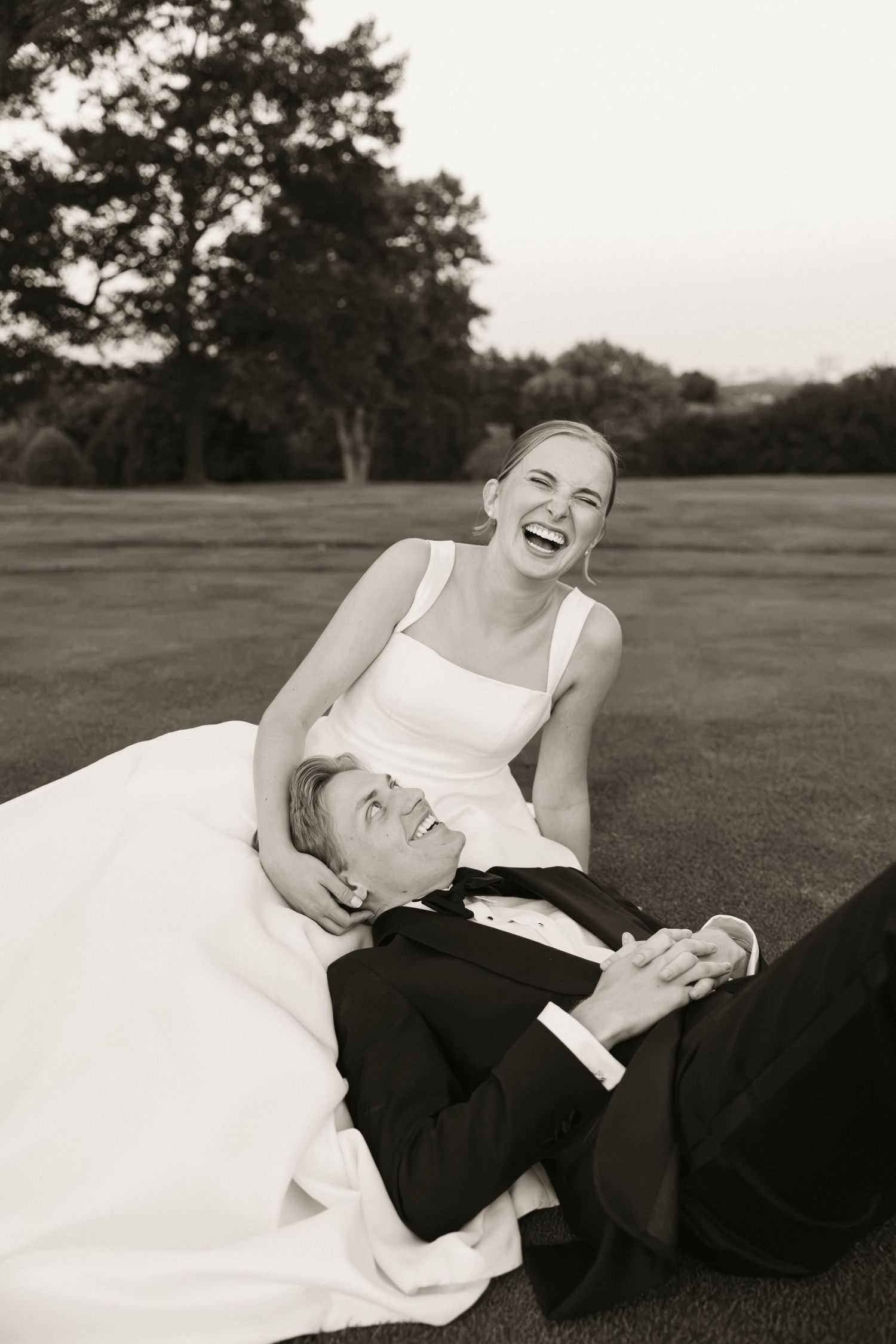 Black and white photo of laughing bride sitting with groom lying in her lap on a golf course at sunset.