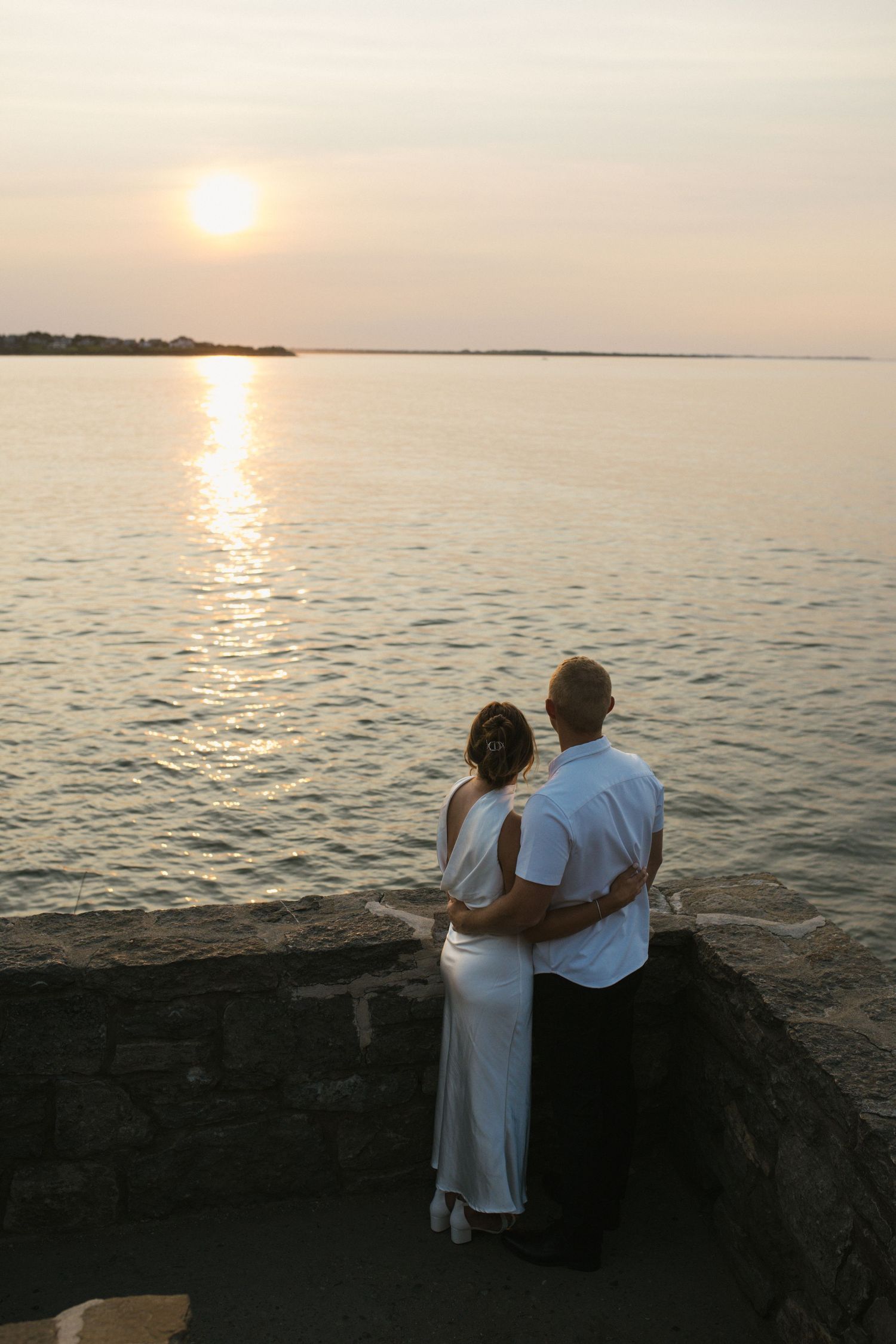 Couple embracing while watching a romantic sunset over calm ocean waters from a stone waterfront wall.