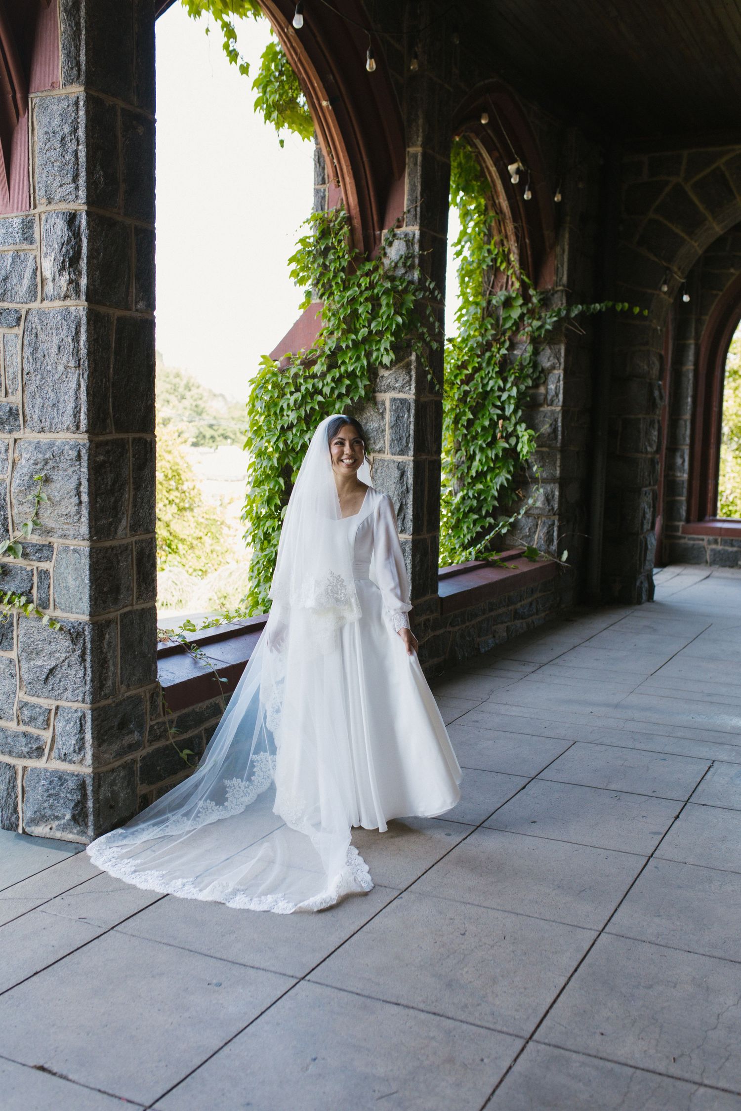 Bride in elegant white gown and cathedral veil poses in ivy-covered stone church archway.