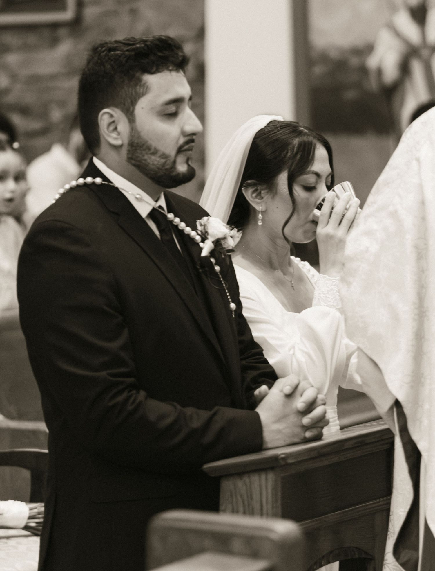 Black and white photo of a bride wiping tears and groom kneeling in prayer during a church wedding ceremony.