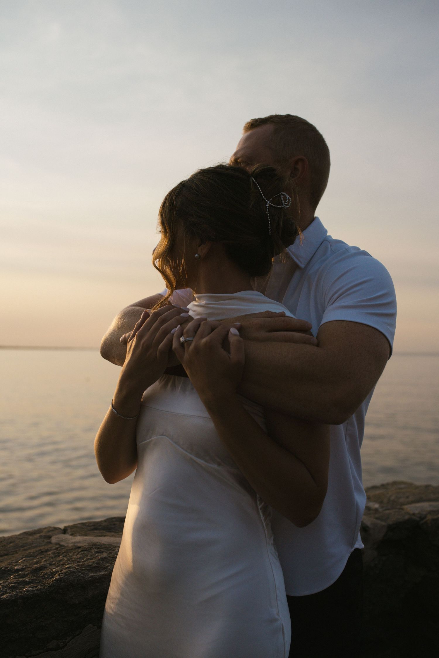 Man tenderly embracing woman from behind on rocky shore during a pastel sunset.