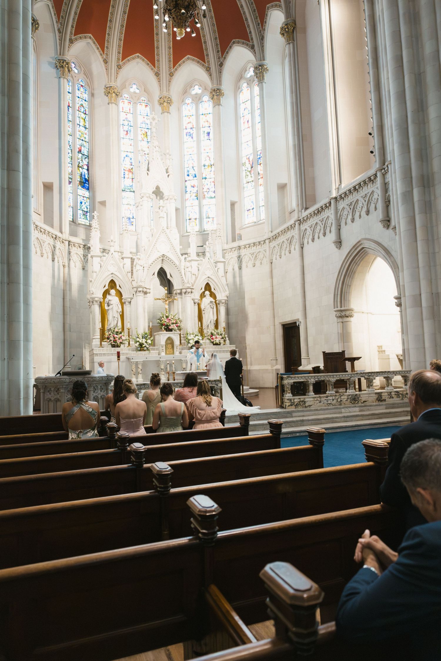 Wedding ceremony in a grand cathedral with stained glass windows, guests seated in wooden pews facing the altar.